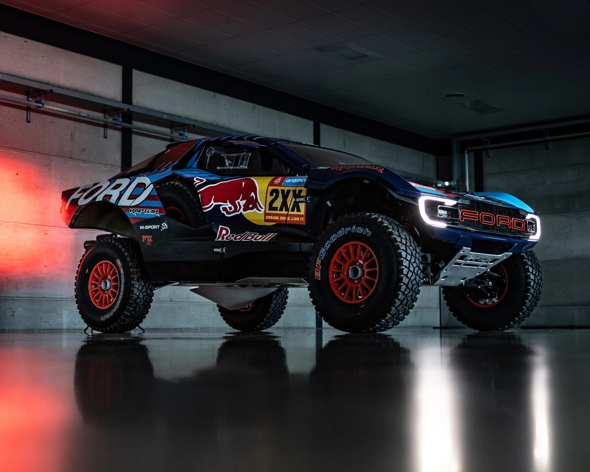 A Ford Raptor off-road racing truck with Red Bull and 2XU sponsorship decals, featuring large BFGoodrich tires with red rims, illuminated LED headlights, and a rugged undercarriage skid plate, parked indoors on a reflective floor with a concrete wall background.