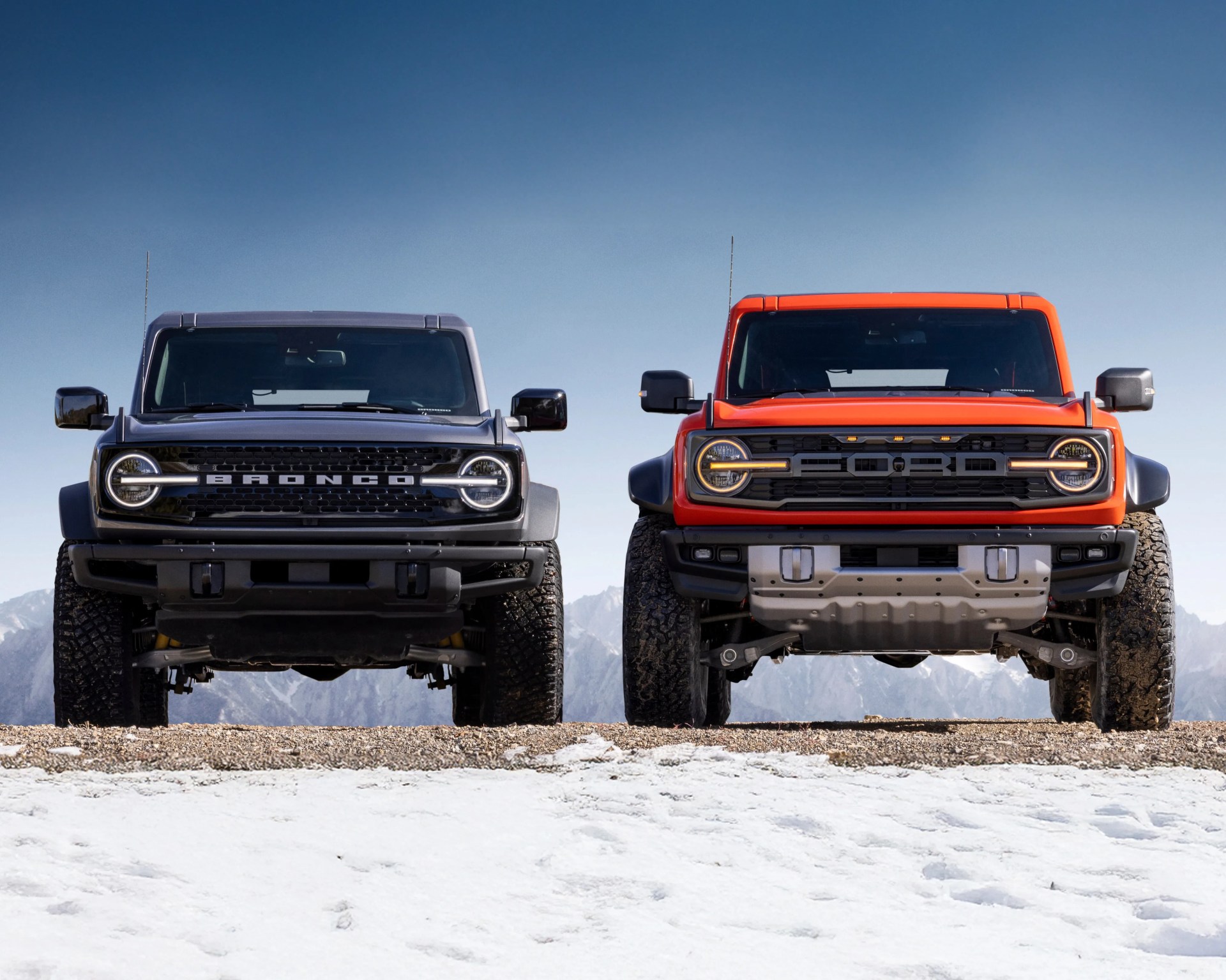 Two Ford Bronco SUVs are parked side by side on a snowy and rocky terrain with mountains in the background. The left vehicle is black with "BRONCO" spelled out on the grille, and the right vehicle is orange with "FORD" spelled out on the grille. Both have round headlights with horizontal light bars crossing through them and large off-road tires. The sky is clear and blue.