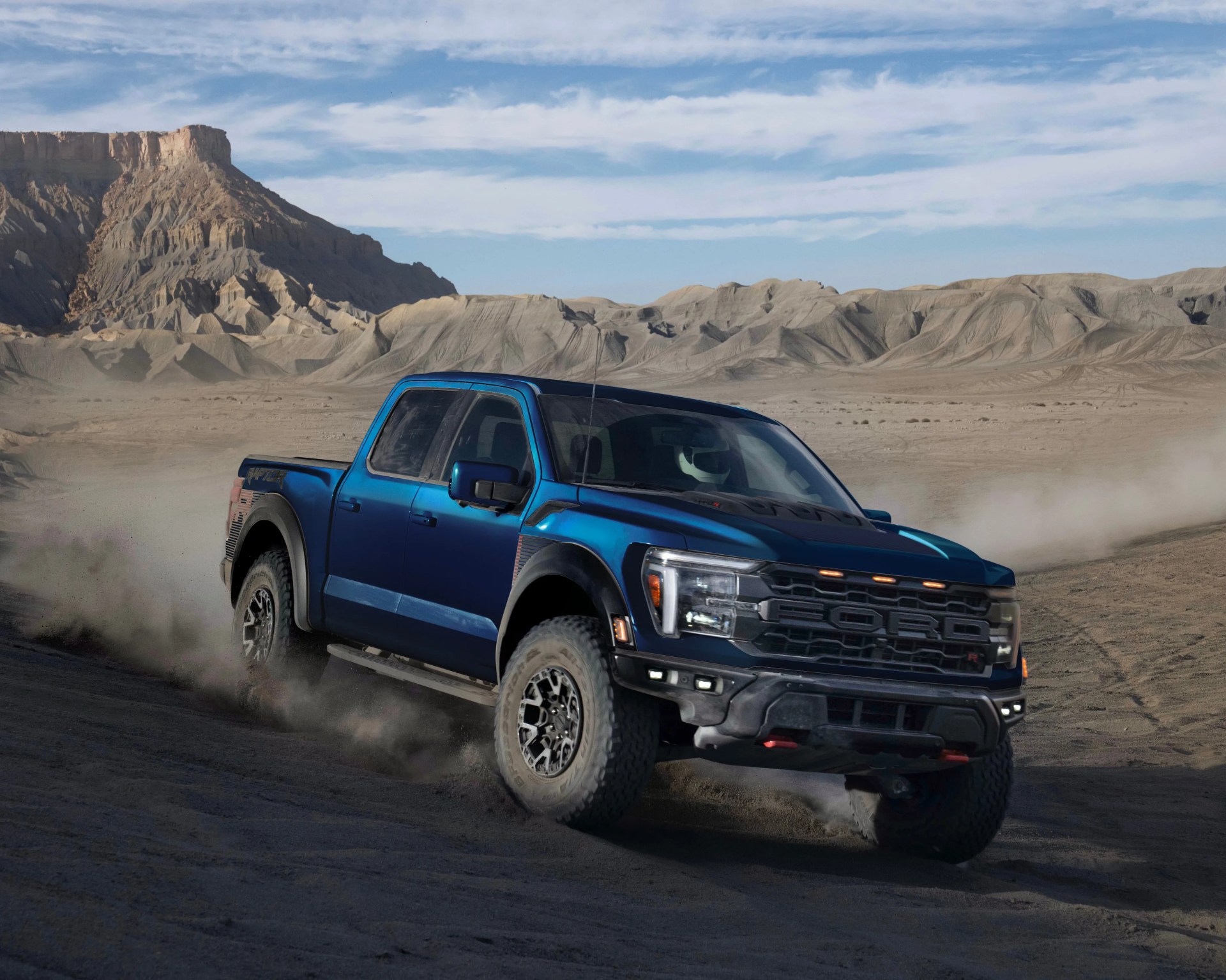 Blue Ford Raptor pickup truck driving off-road on a dusty desert terrain with rugged hills and a partly cloudy sky in the background.