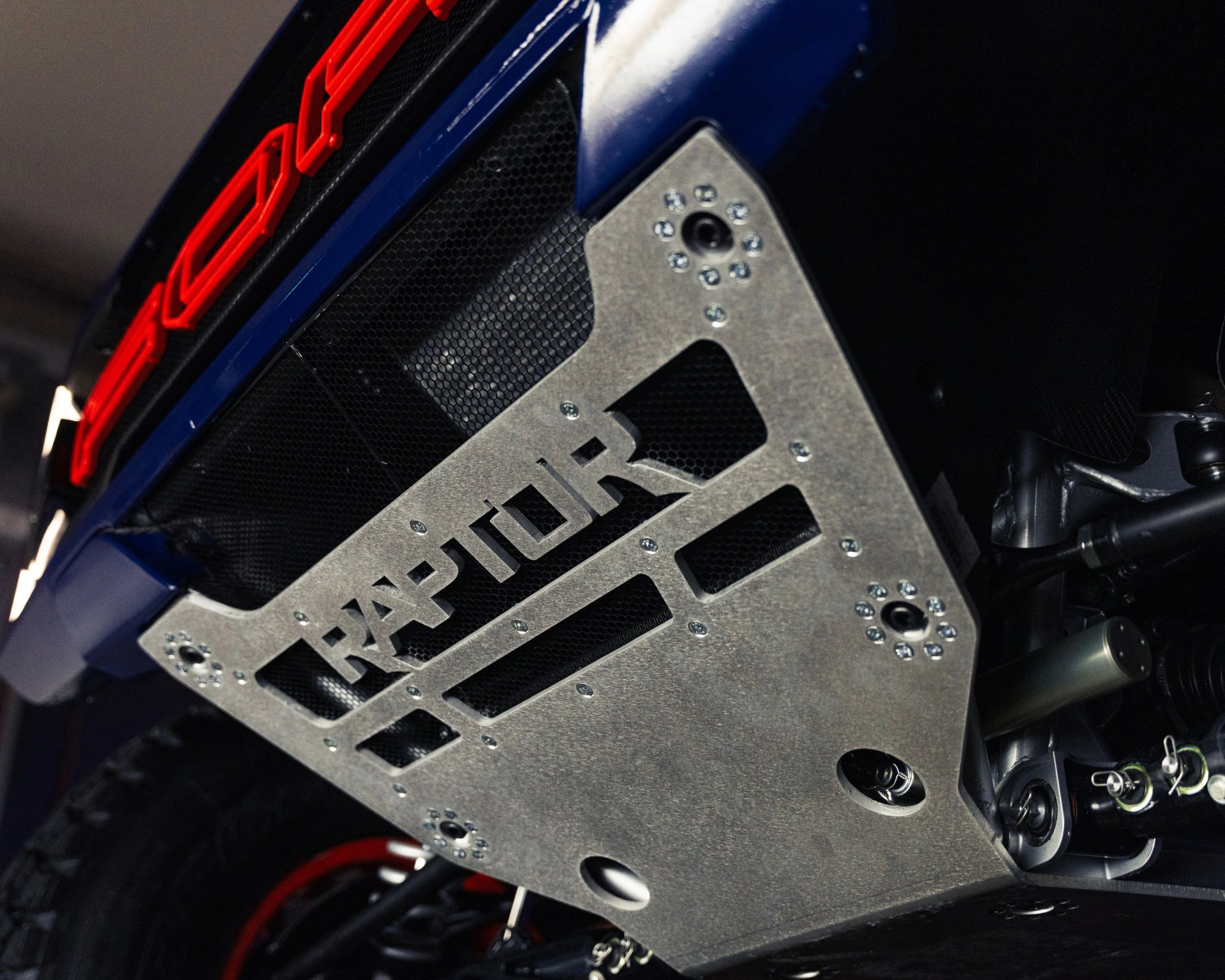 Underside view of a vehicle featuring a metal skid plate with the word "RAPTOR" cut out, secured with multiple bolts. The vehicle has a blue body with a red "FORD" emblem visible above the skid plate. The image also shows part of the suspension system and a tire with a red rim.
