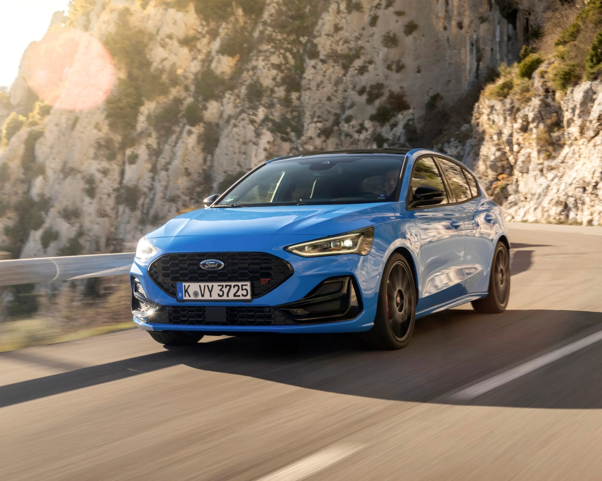 Blue Ford Focus ST hatchback driving on a winding mountain road with rocky cliffs in the background, featuring black wheels and a black front grille.