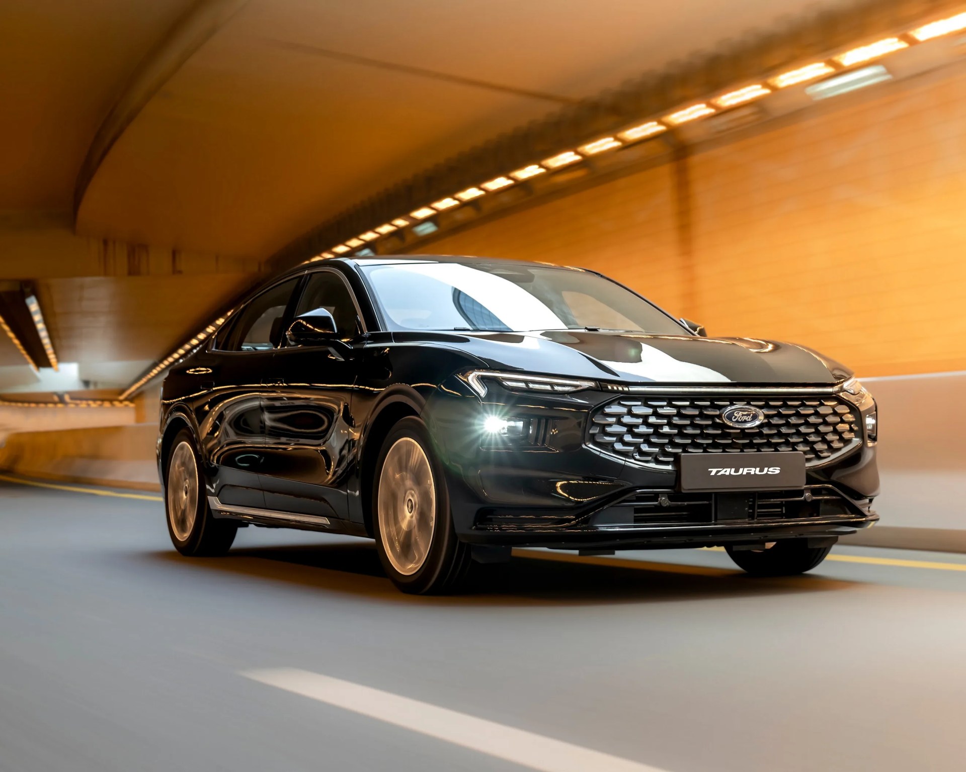 Black Ford Taurus sedan driving through a tunnel with illuminated orange walls, featuring a modern front grille and headlights turned on.
