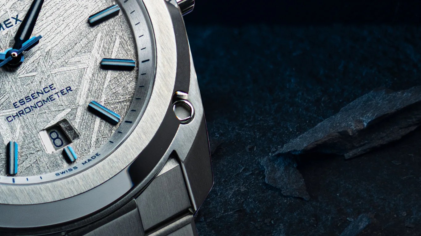 Close-up of a silver formex wristwatch with a textured dial featuring blue hour markers and hands. The dial includes the text "ESSENCE CHRONOMETER" and "SWISS MADE," with a date window displaying the number 8. The watch has a stainless steel bezel and bracelet, set against a dark, textured background.