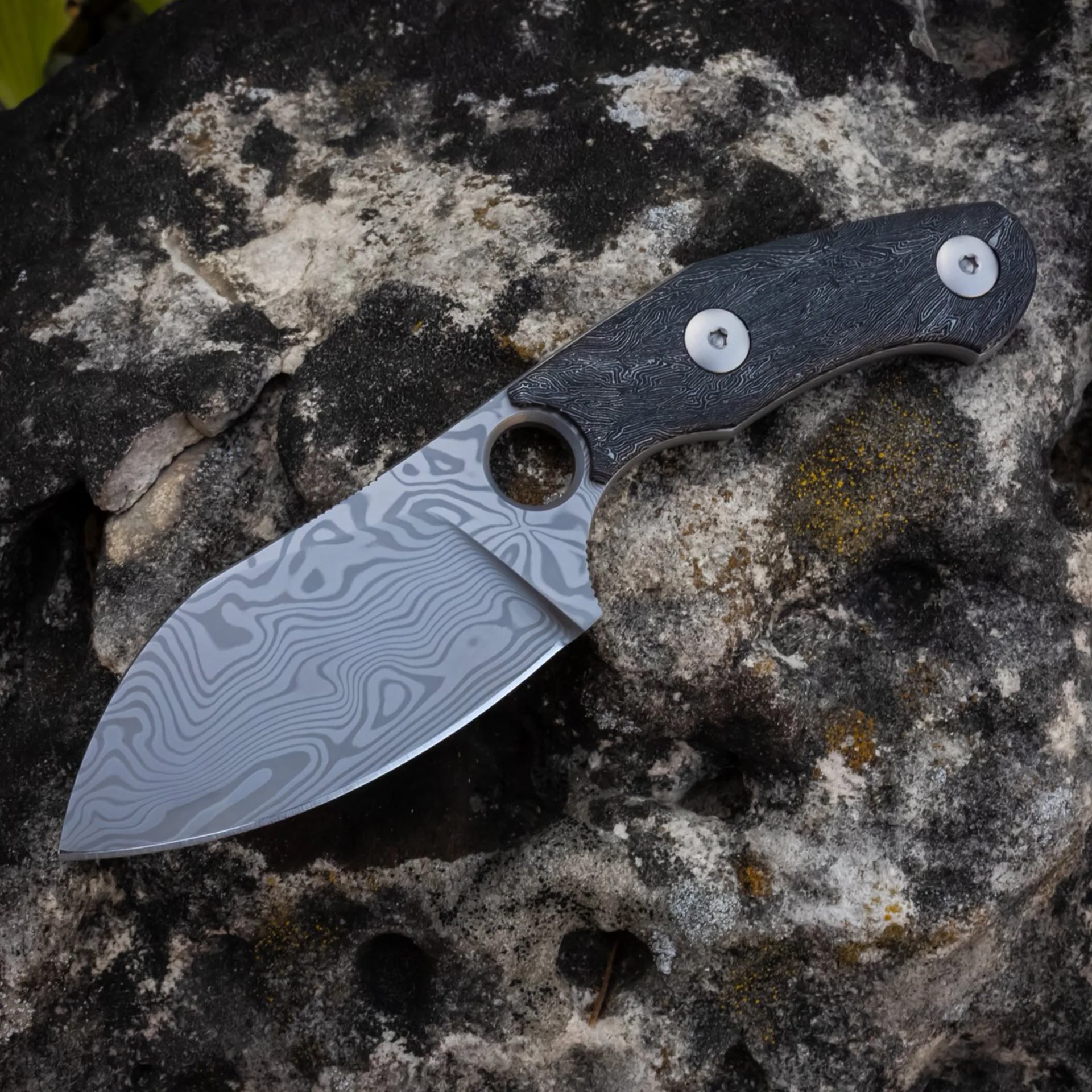 A fixed-blade knife with a patterned Damascus steel blade and a dark textured handle secured by two silver screws, resting on a rough, mottled rock surface. The blade has a distinctive wavy pattern and a circular hole near the handle.
