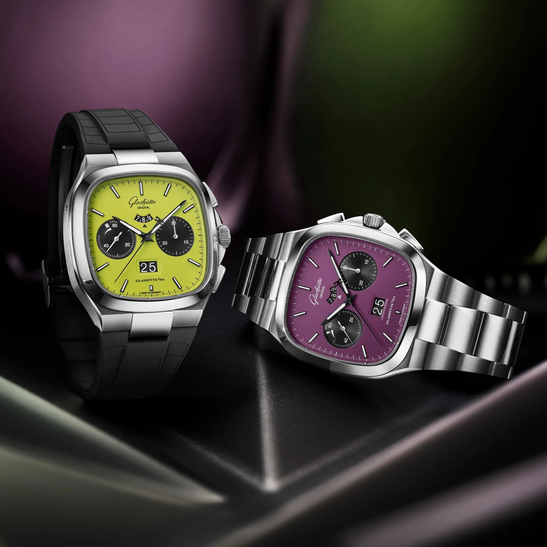 Two Glashütte Original wristwatches with square stainless steel cases and chronograph features. The watch on the left has a bright green dial, black subdials, a black rubber strap, and a large date display at 6 o'clock. The watch on the right has a purple dial, black subdials, a stainless steel bracelet, and a large date display at 6 o'clock. Both watches have silver hour markers and hands, with the brand name "Glashütte Original" visible below the 12 o'clock position.