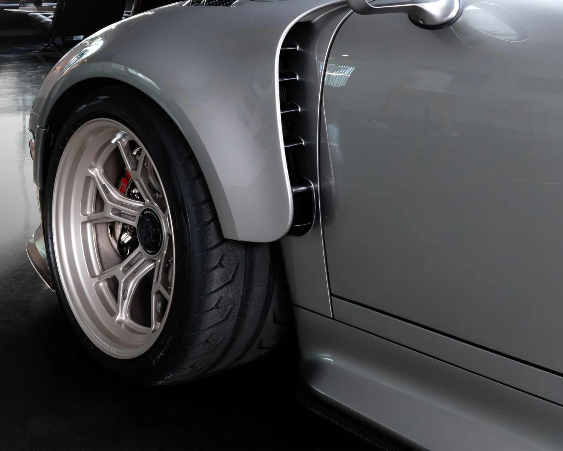 Close-up of a silver sports car's rear wheel and fender, featuring a wide tire with a detailed tread pattern, a multi-spoke alloy rim, and a visible red brake caliper. The car's body has a smooth, glossy finish with an air vent behind the wheel and a side mirror above.
