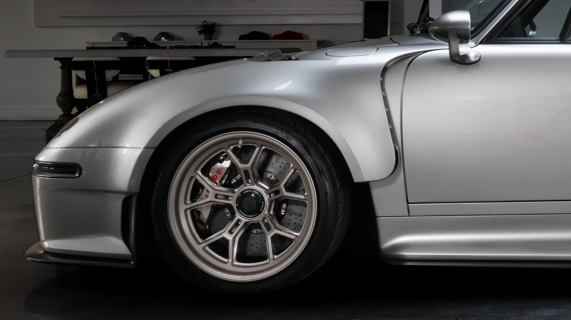 Close-up of the front left side of a silver sports car, showcasing a detailed alloy wheel with a visible Brembo brake caliper and drilled brake disc. The car features a smooth, aerodynamic fender and side mirror, with a subtle front splitter visible at the bottom left. The background includes a table and chairs in a dimly lit indoor setting.