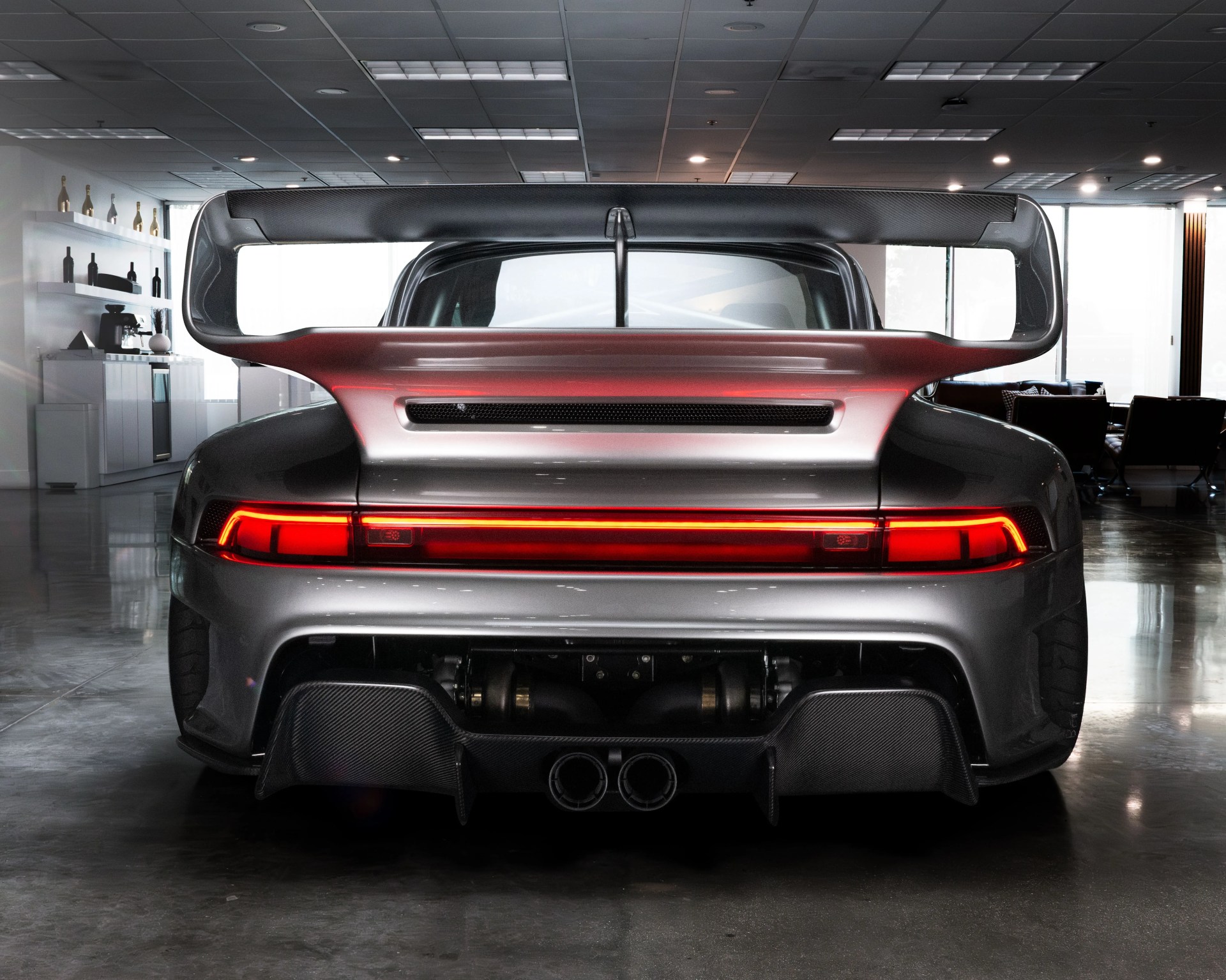 Rear view of a silver sports car with a large carbon fiber rear wing, a continuous red LED tail light strip, dual exhaust pipes, and a carbon fiber diffuser, parked indoors on a polished concrete floor.