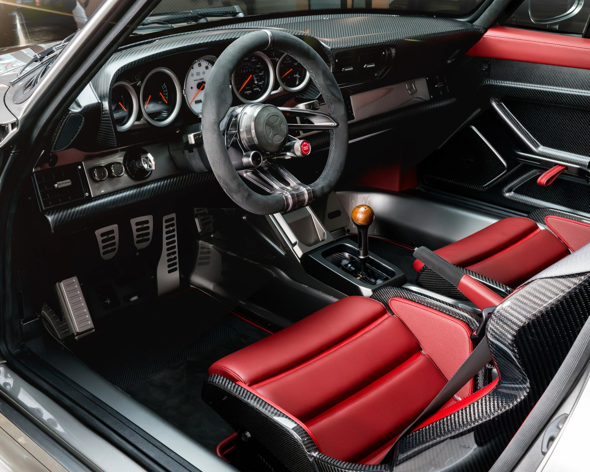 Sporty car interior featuring red leather bucket seats with carbon fiber accents, a suede-wrapped steering wheel with metallic spokes, a wooden gear shift knob, and a dashboard with multiple round gauges and carbon fiber trim.