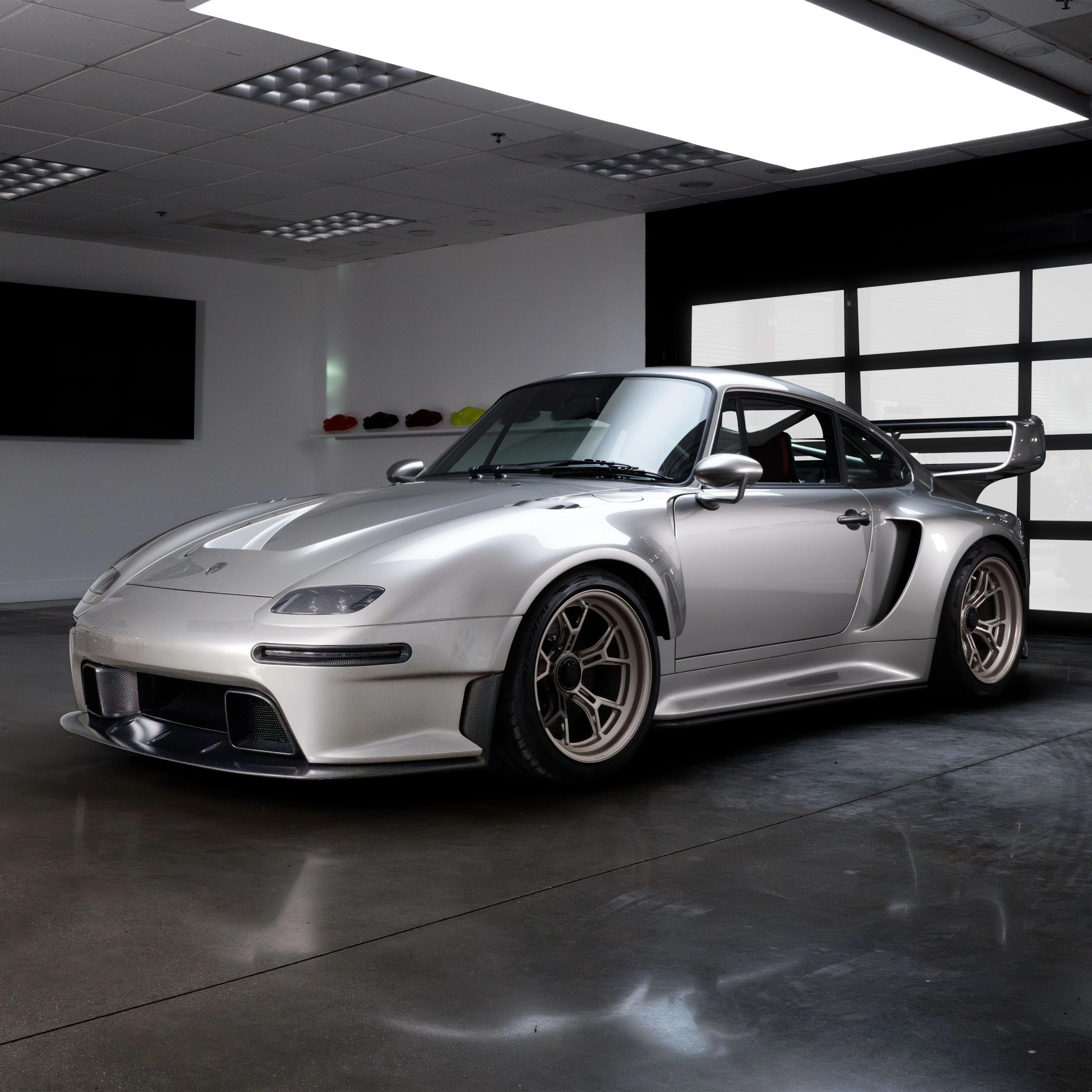 Silver Porsche 911 GT2 RS with wide body kit and large rear wing, parked indoors on a polished concrete floor, under bright ceiling lights. The car features bronze multi-spoke wheels and aerodynamic front splitter.
