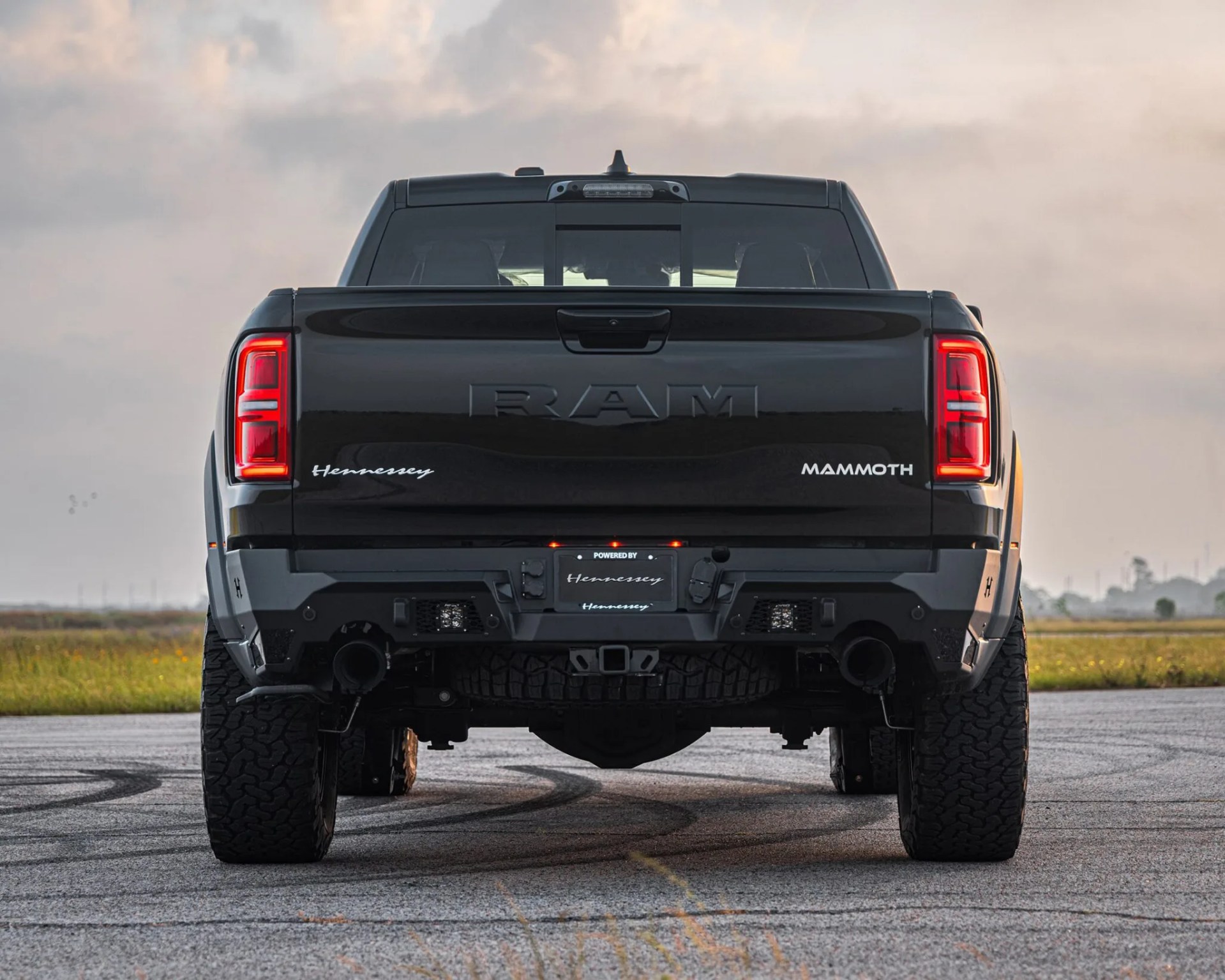 Rear view of a black RAM pickup truck with "Hennessey" and "Mammoth" badges on the tailgate, featuring illuminated red taillights, a rugged rear bumper, and large off-road tires, parked on a paved surface with a grassy field and cloudy sky in the background.
