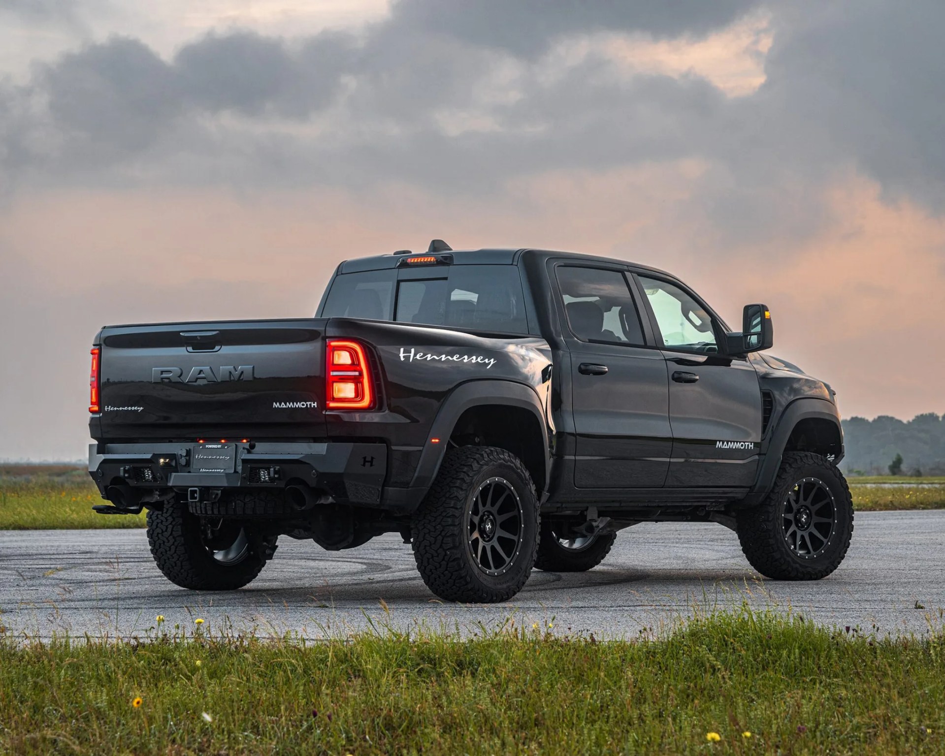 Black RAM Mammoth pickup truck with Hennessey branding, featuring large off-road tires and a lifted suspension, parked on a paved surface with grass in the foreground and a cloudy sky at sunset in the background.