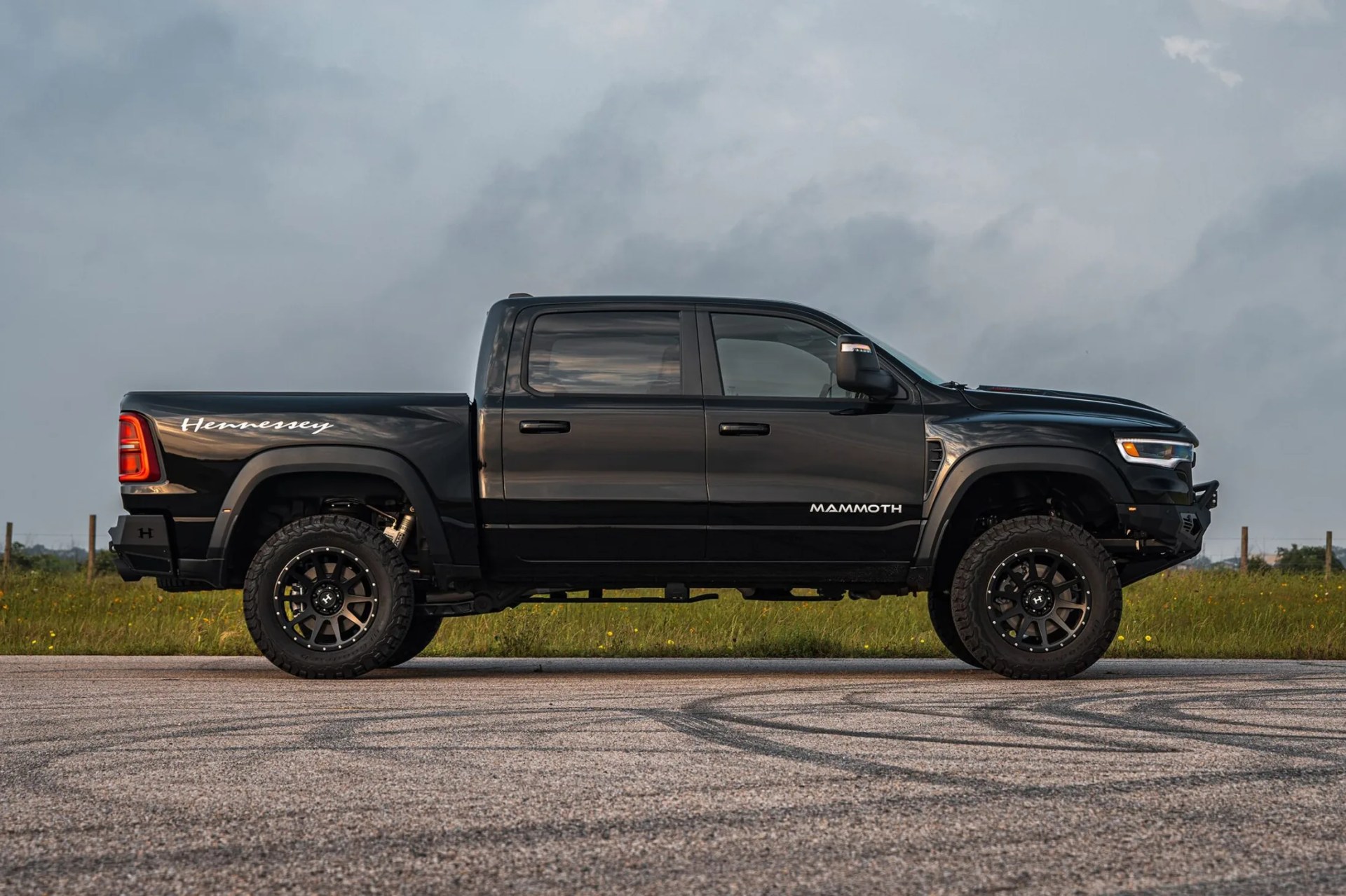 Black Hennessey Mammoth pickup truck with off-road tires, black alloy wheels, and tinted windows, parked on a paved surface with grass and a cloudy sky in the background.
