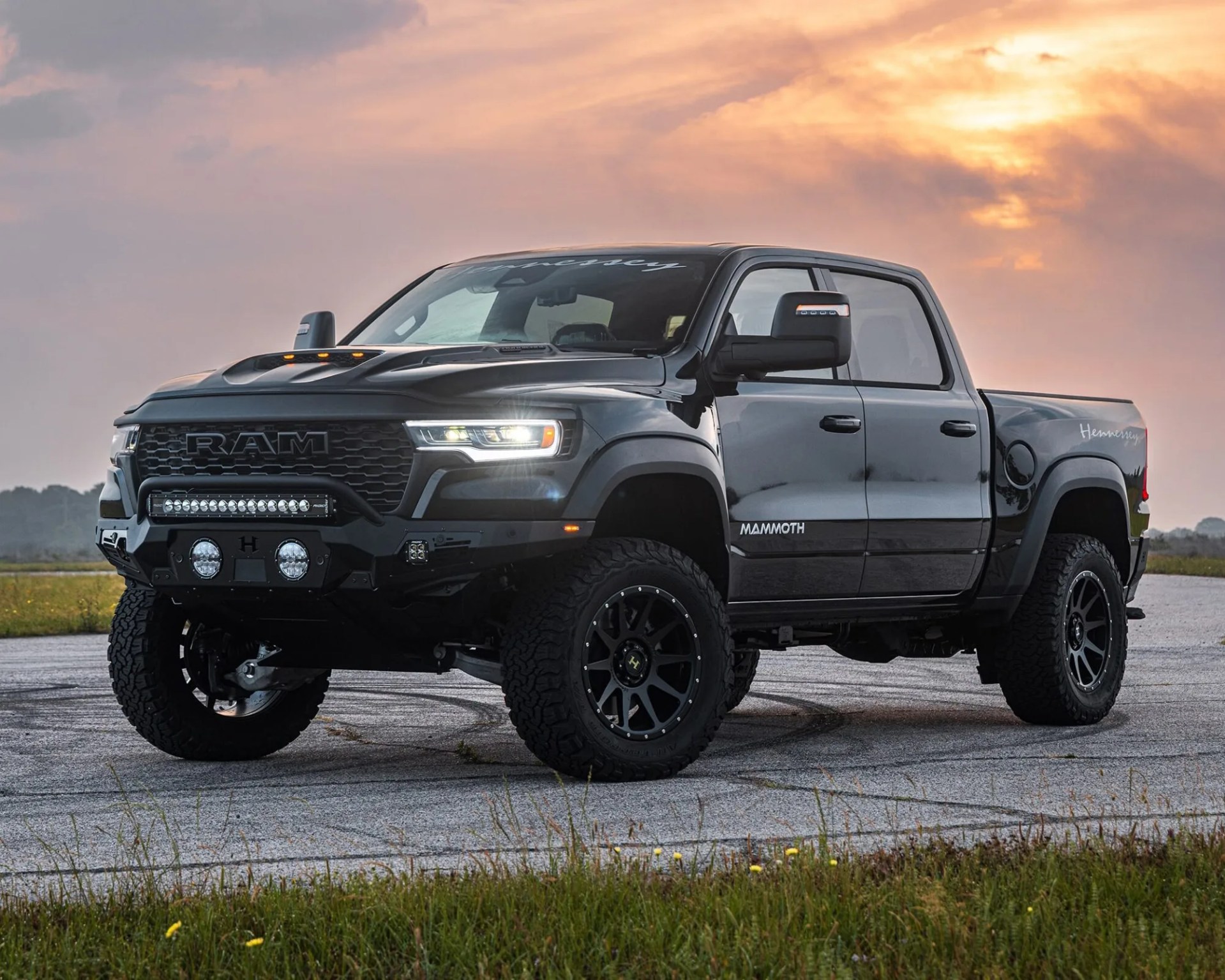 Black RAM pickup truck with "Mammoth" and "Hennessey" branding, featuring off-road tires, a rugged front bumper with additional lights, and a hood scoop, parked on a paved surface at sunset.