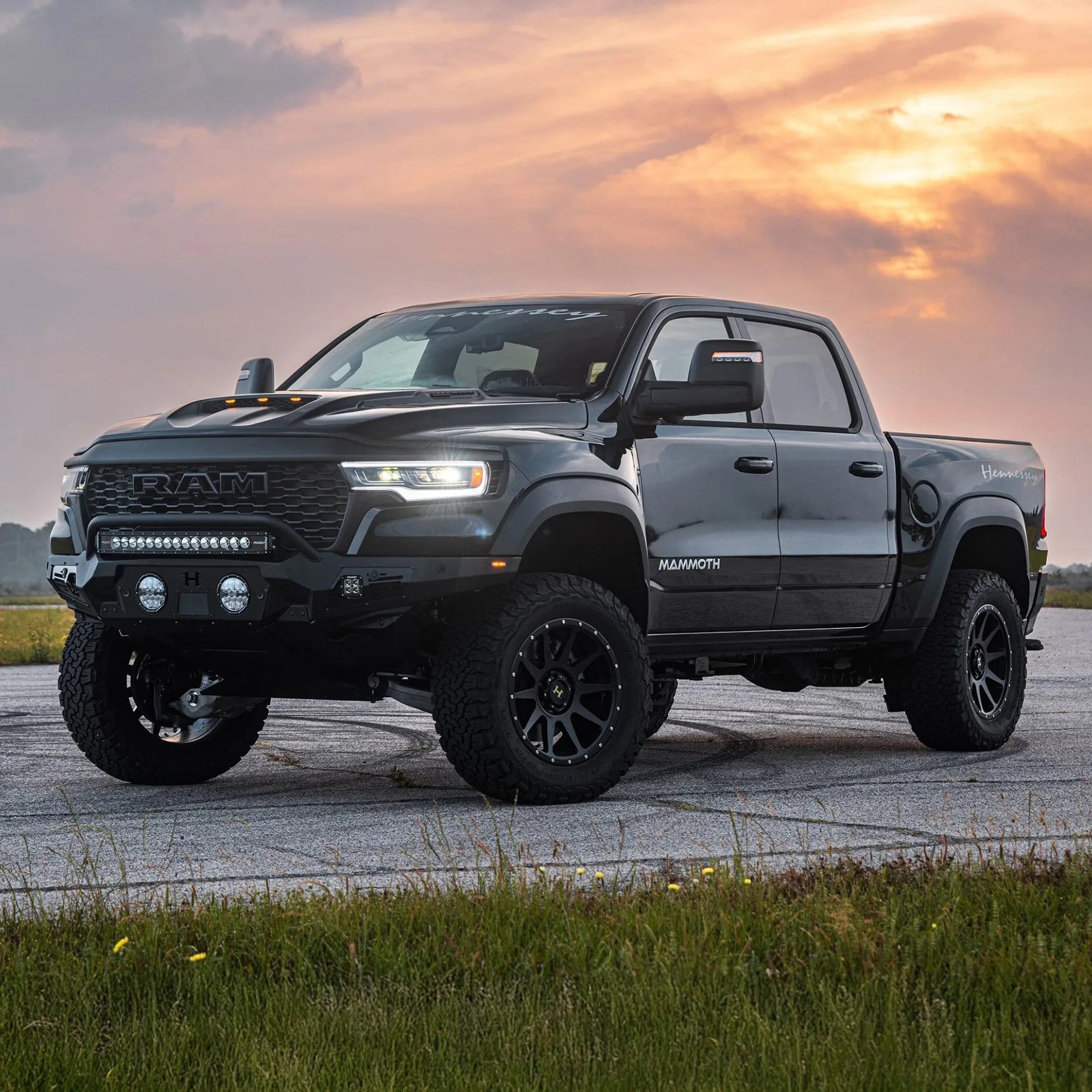 Black RAM pickup truck with off-road tires and a reinforced front bumper, parked on a paved surface with grass in the foreground and a sunset sky in the background. The truck has "MAMMOTH" written on the side and "Hennessey" on the rear side panel.