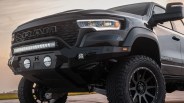 Front side view of a black and white RAM Mammoth pickup truck with large off-road tires, a heavy-duty black front bumper equipped with multiple LED lights, and a prominent RAM grille. The truck has a rugged, lifted stance suitable for off-road driving.