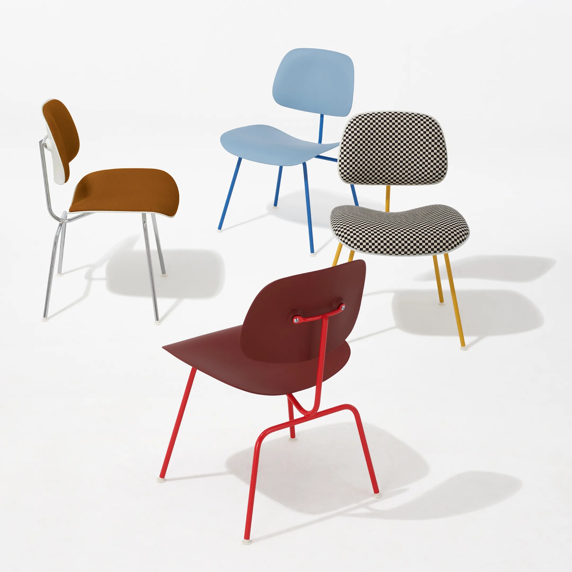 Four modern herman miller eames chairs with distinct designs and colors are arranged on a white background. One chair has a brown cushioned seat and back with chrome legs. Another chair is light blue with a smooth seat and back and blue legs. A third chair features a black-and-white checkered cushioned seat and back with yellow legs. The fourth chair has a maroon seat and back with red metal legs and a unique support structure on the backrest.