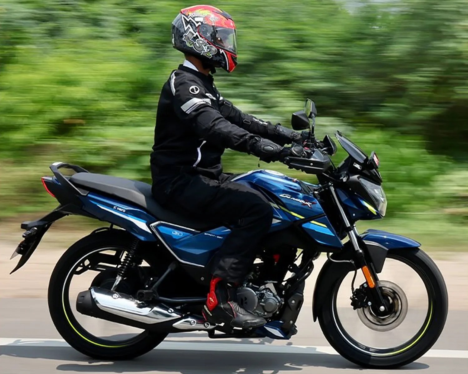 A person wearing a black riding suit, black gloves, and a red and black helmet is riding a blue Hero motorcycle on a road with green foliage in the background. The motorcycle has yellow and white accents and features a chrome exhaust pipe.