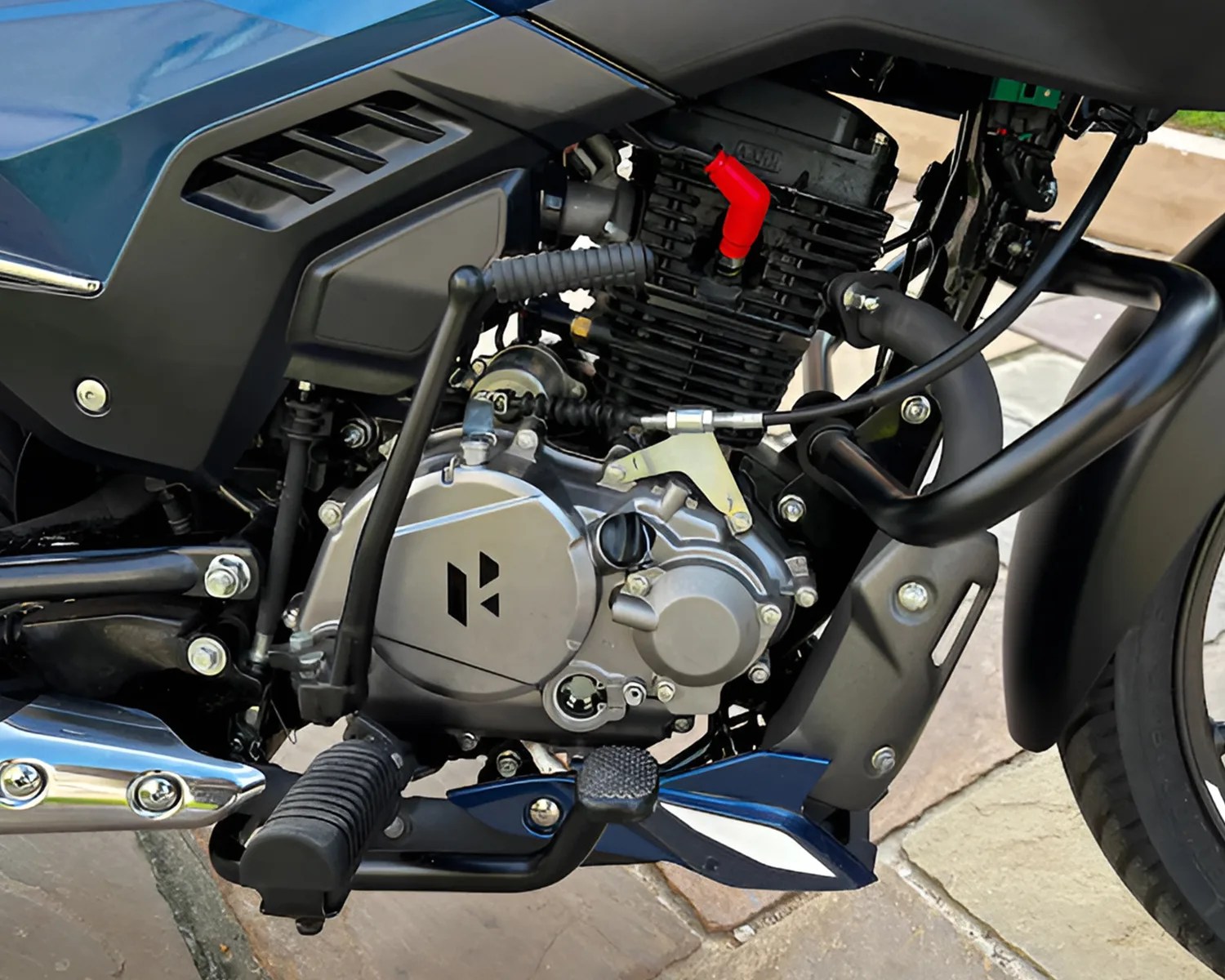 Close-up of a motorcycle engine and surrounding components, including the foot pedal, gear shift lever, exhaust pipe, and part of the rear wheel. The engine casing is silver with a black logo, and there is a red spark plug cap visible on the black engine block. The frame and other parts around the engine are mostly black with some blue accents. The motorcycle is parked on a stone-paved surface.