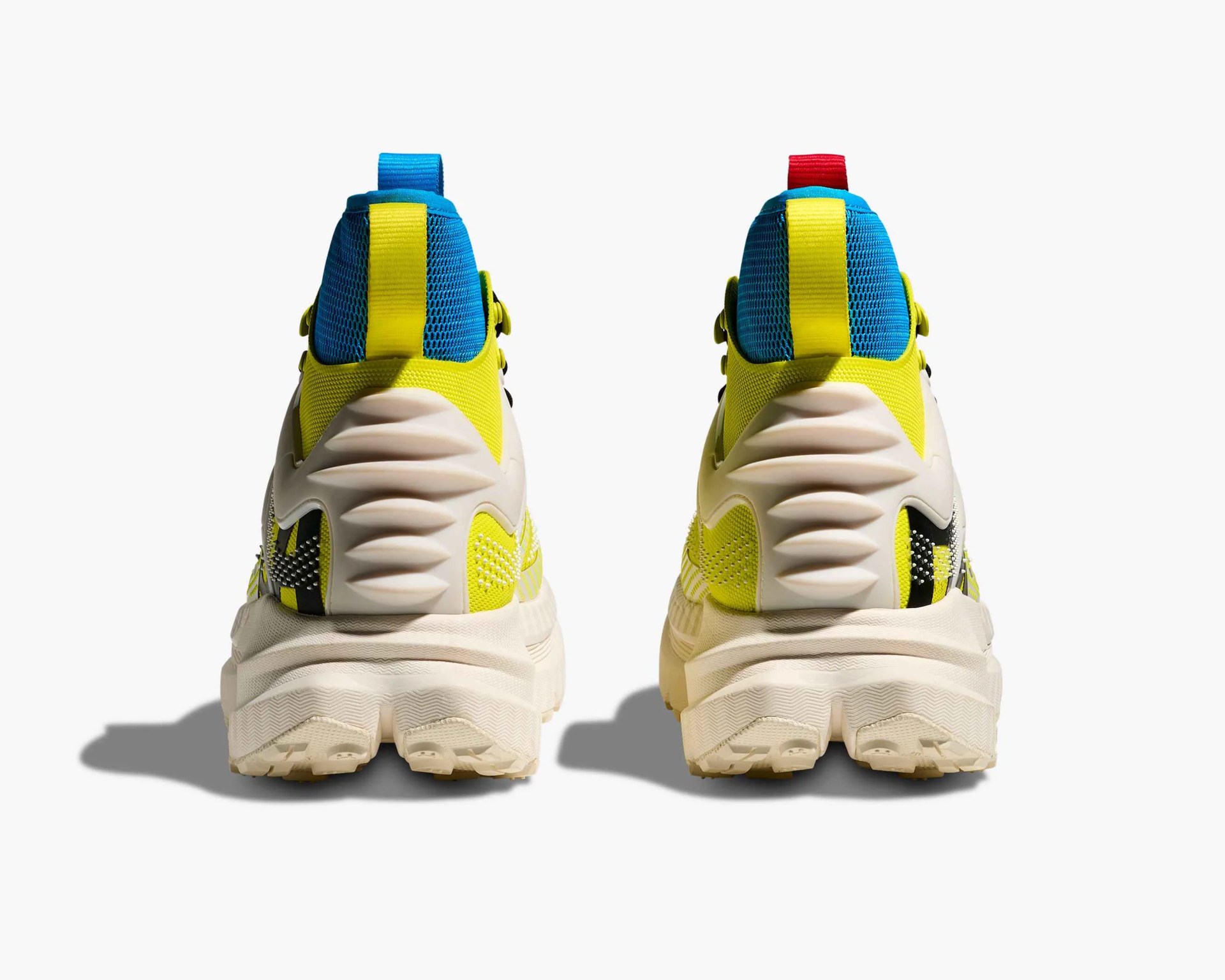 Pair of athletic shoes shown from the back, featuring a chunky off-white sole with a textured pattern. The upper part of the shoes is primarily bright yellow with blue mesh around the ankle area. Each shoe has a vertical pull tab on the heel, one in blue and the other in red. The heel area also includes a sculpted off-white plastic overlay with a wave-like design.