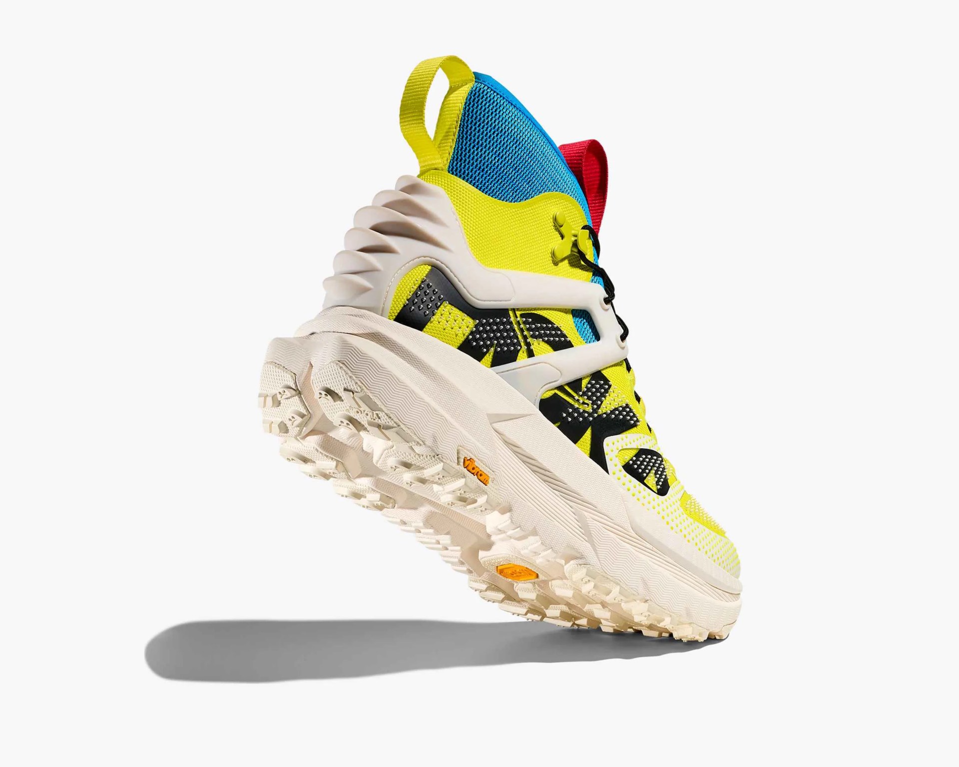 High-top athletic shoe with a chunky, textured off-white sole featuring deep treads and Vibram branding. The upper part of the shoe is vibrant yellow with black geometric patterns and small white dots. It has a blue mesh section near the ankle, a red pull tab at the back, and a yellow pull tab at the heel. The shoe also includes a white supportive cage structure around the midfoot and black laces. The heel has a sculpted, ribbed design for added style and support.