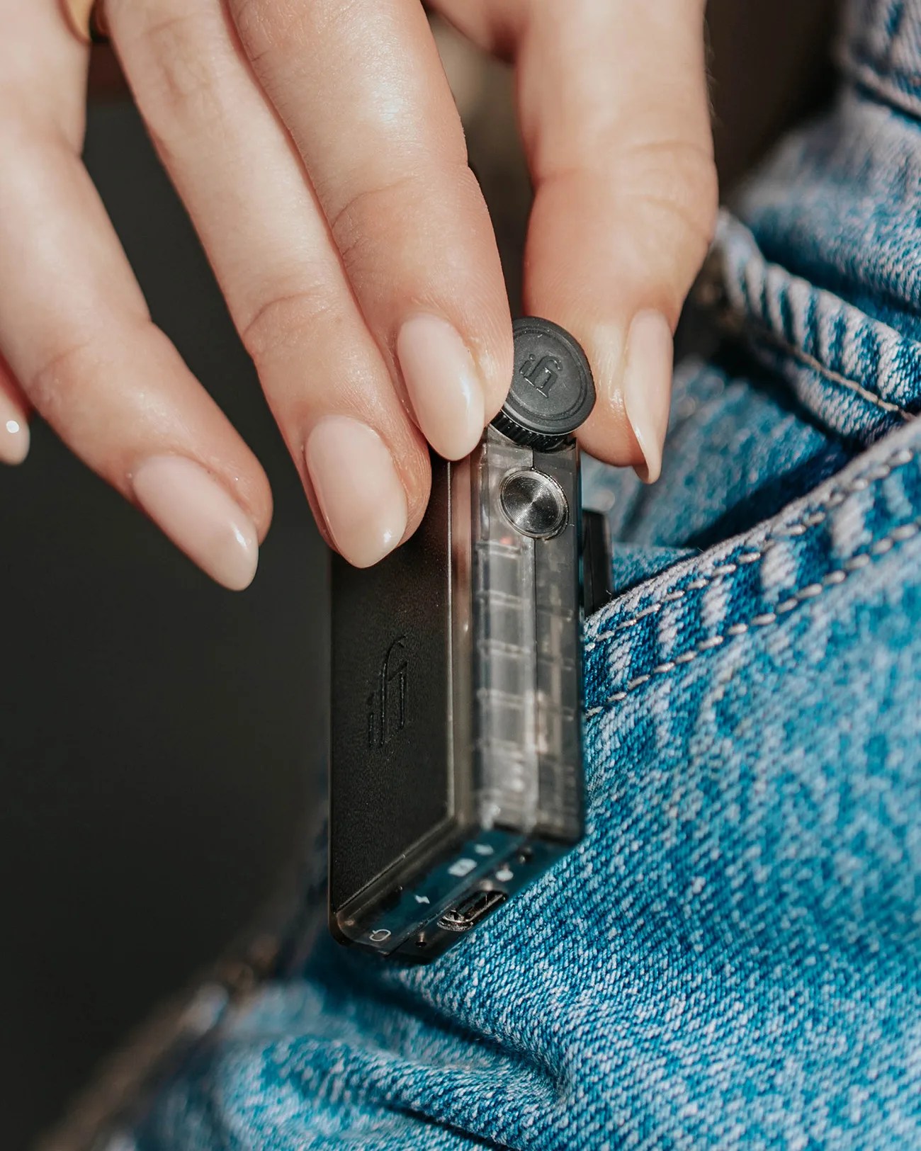 A hand with neatly manicured nails is holding a small, rectangular black electronic device with a transparent side and a circular button or knob on top. The device is being clipped onto the pocket of blue denim jeans.