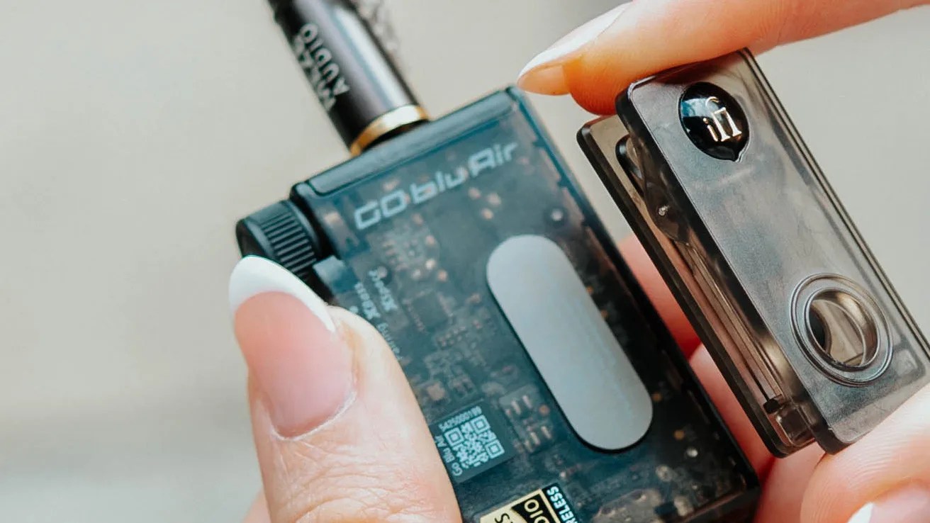 A close-up of a hand holding a small electronic device labeled "Goblin Air" with a transparent casing showing internal circuitry, and a detachable component being removed or attached. The device appears to be a compact vaping or pod system.