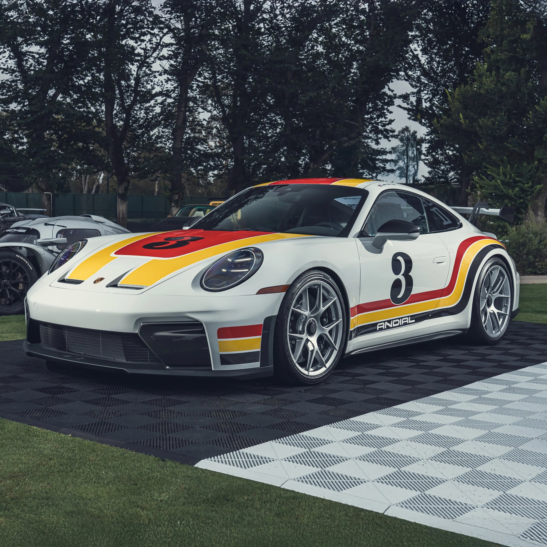 White Porsche sports car with red, yellow, and black racing stripes and the number 3 on the hood and door, parked on a checkered black and white mat outdoors with trees in the background.