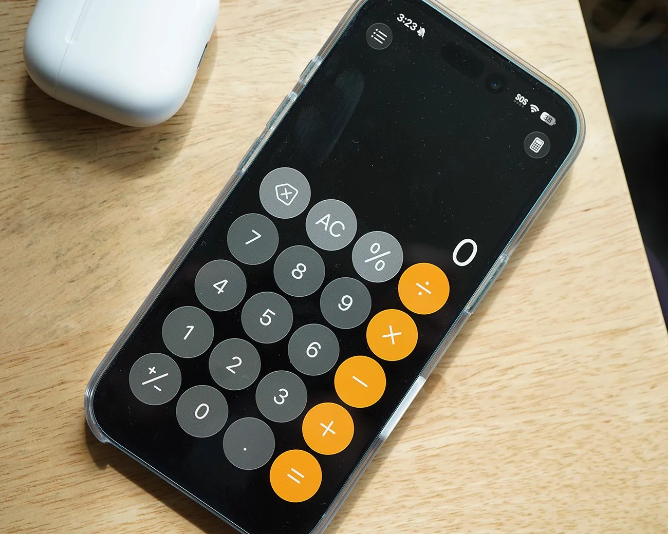 A smartphone with a clear protective case displaying a calculator app on its screen, placed on a light wooden surface. The calculator app shows a zero on the display and has gray number buttons and orange operation buttons. A white wireless earbud case is partially visible in the upper left corner.