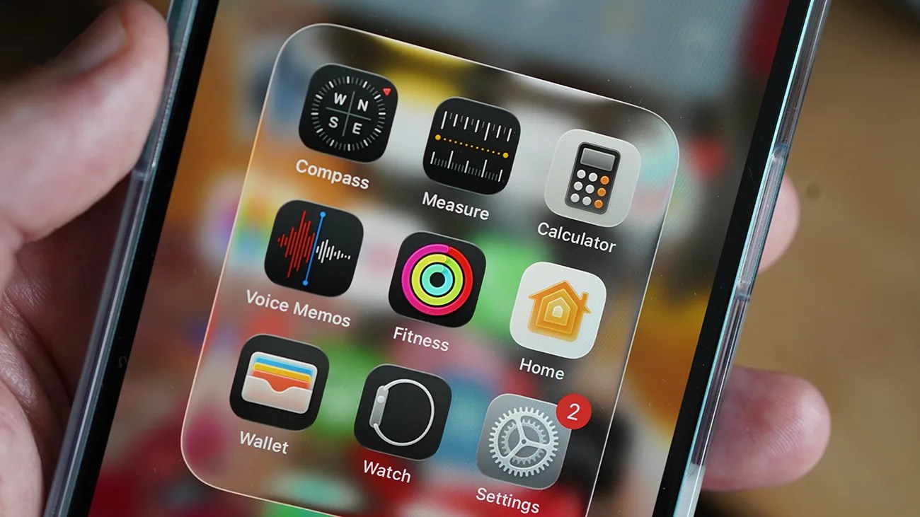 A smartphone screen displaying a folder with nine app icons: Compass, Measure, Calculator, Voice Memos, Fitness, Home, Wallet, Watch, and Settings with a red notification badge showing the number 2. The phone is held in a hand.