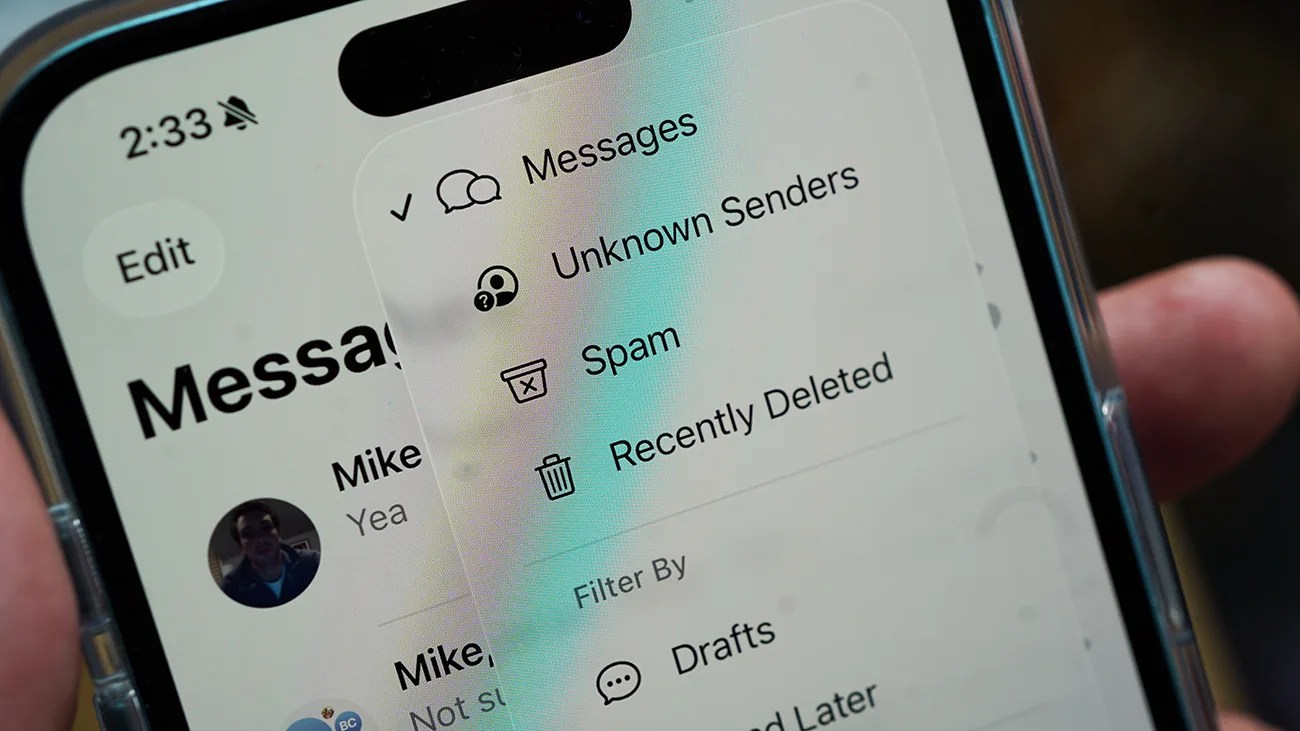 Close-up of a smartphone screen displaying the Messages app with a filter menu open. The menu options include Messages, Unknown Senders, Spam, Recently Deleted, and Drafts. The time on the phone is 2:33, and the phone is in silent mode. A person's hand is holding the phone.
