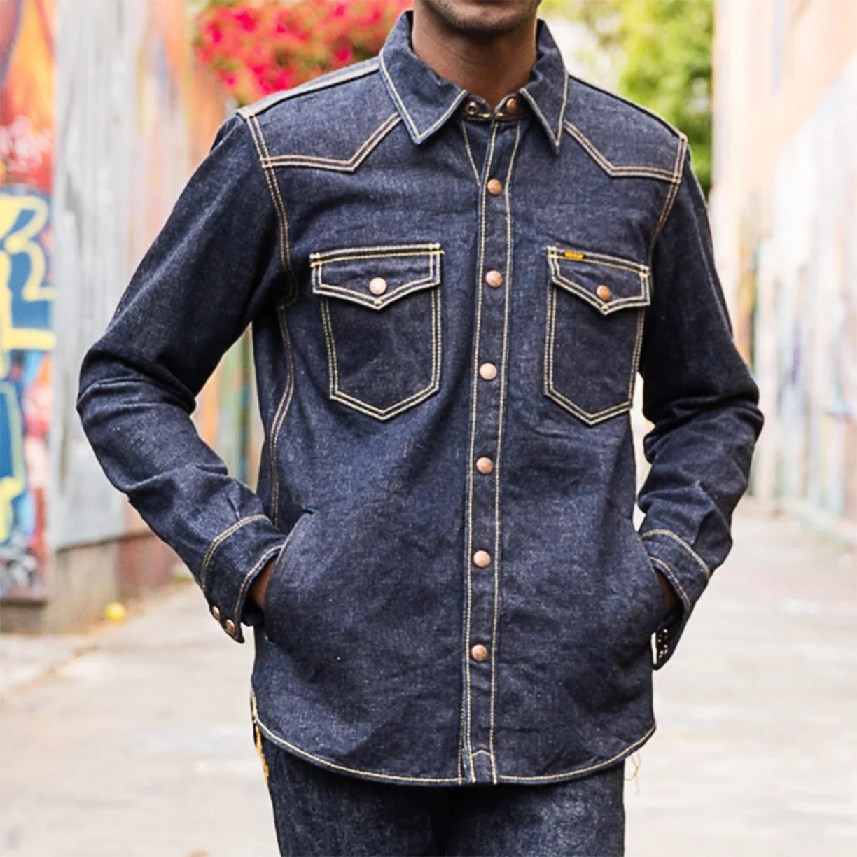 Dark blue denim jacket with contrast yellow stitching, featuring two front flap pockets with buttons and copper-colored snap buttons down the front. The jacket has a classic collar and the person wearing it has their hands in the side pockets.