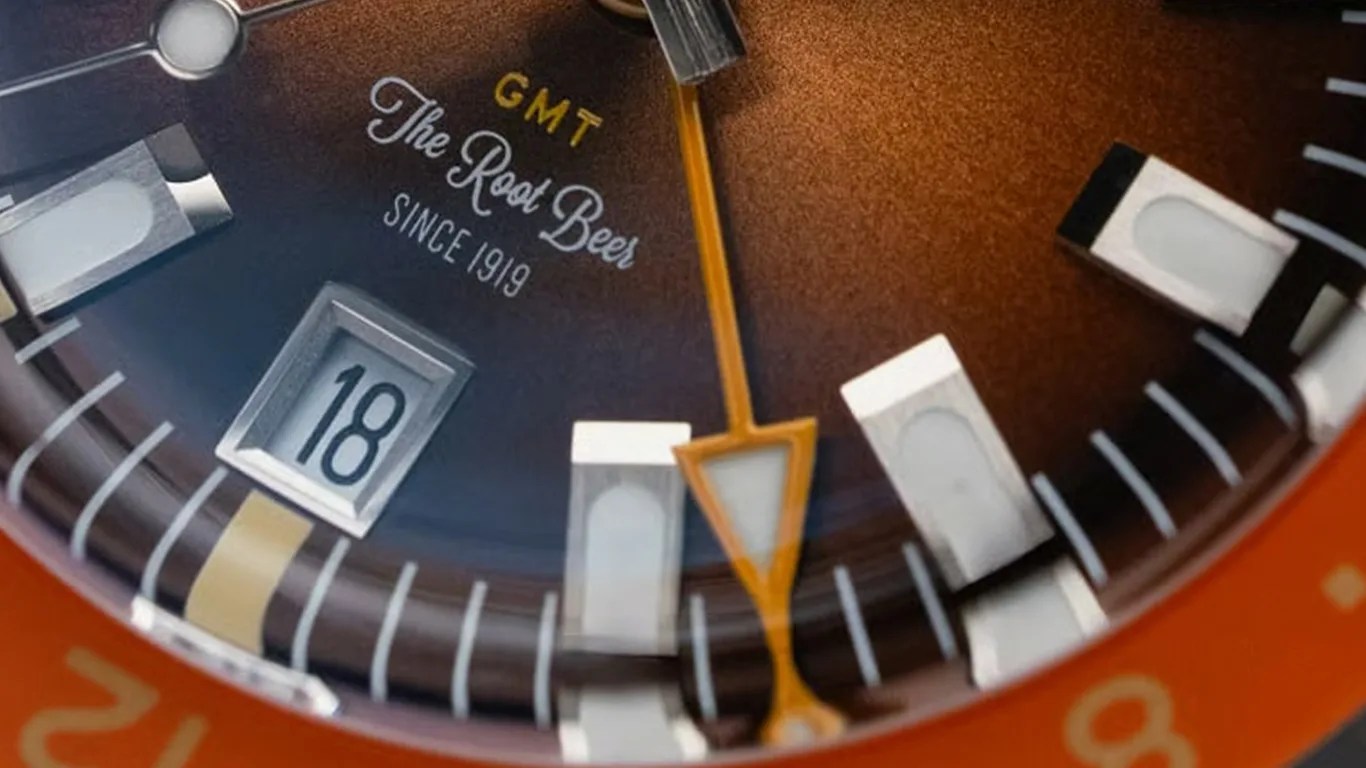 Close-up of a watch face with a brown gradient dial, featuring the text "GMT The Root Beer Since 1919." The date window shows the number 18, and the watch has rectangular hour markers with white luminescent centers. The GMT hand is orange with a triangular tip. The outer bezel is orange with yellow markings.