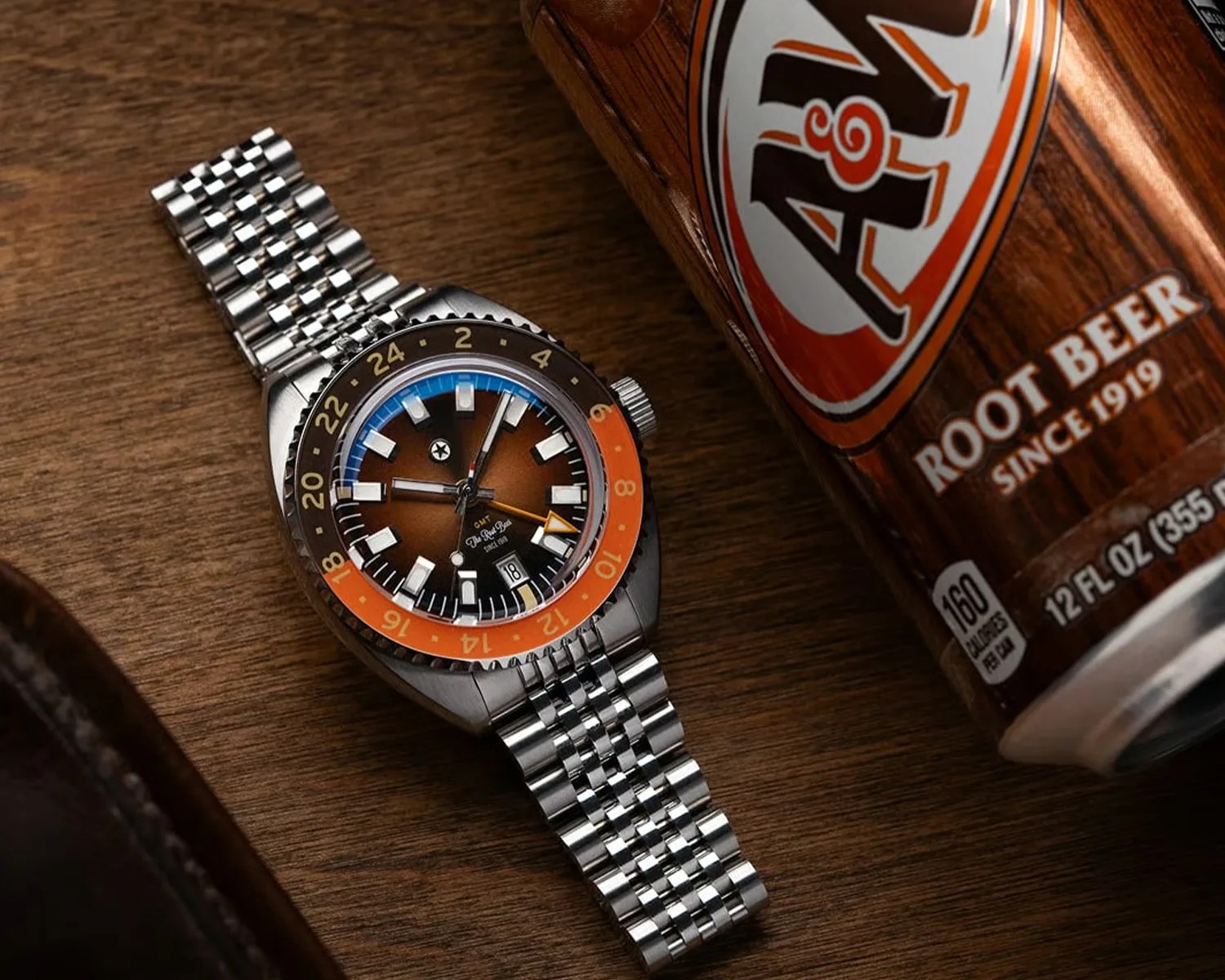 A stainless steel wristwatch with a multi-link bracelet, featuring a brown gradient dial and a bi-color bezel in black and orange with 24-hour markings. The watch has bold white hour markers, a date window at 6 o'clock, and an orange GMT hand. It is placed on a wooden surface next to a can of A&W Root Beer.
