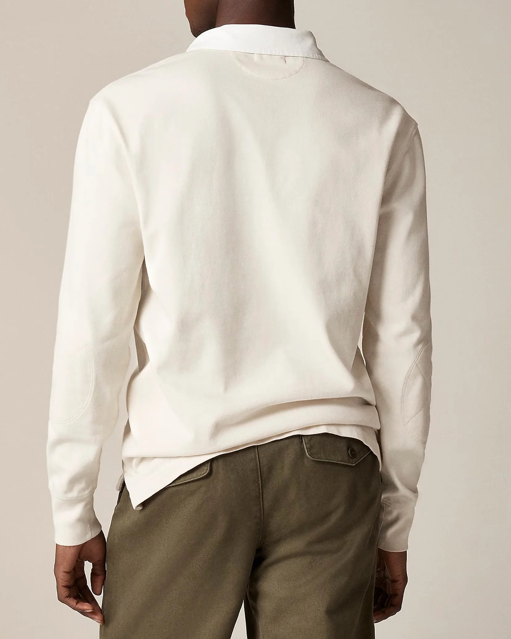 Back view of a person wearing a long-sleeve, off-white polo shirt with elbow patches, paired with olive green pants featuring buttoned back pockets.