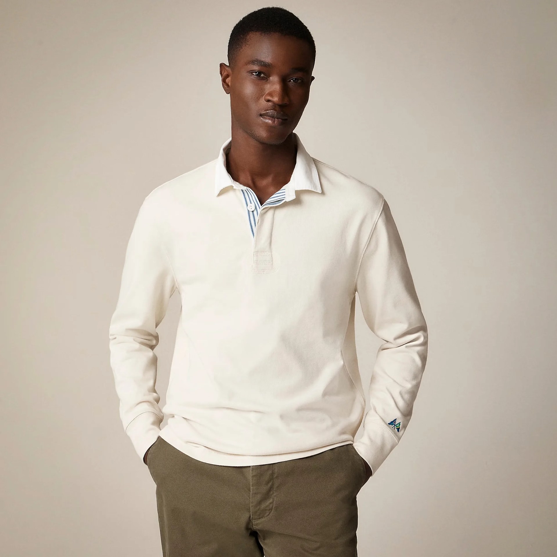 Man wearing a long-sleeve, off-white polo shirt with a collar and a blue and white striped inner placket, paired with olive green pants. The shirt has a small embroidered detail on the left sleeve cuff.