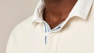 Close-up of a person wearing a cream-colored collared shirt with a white button and a blue and white striped inner placket detail. The collar is slightly open, revealing the neck.