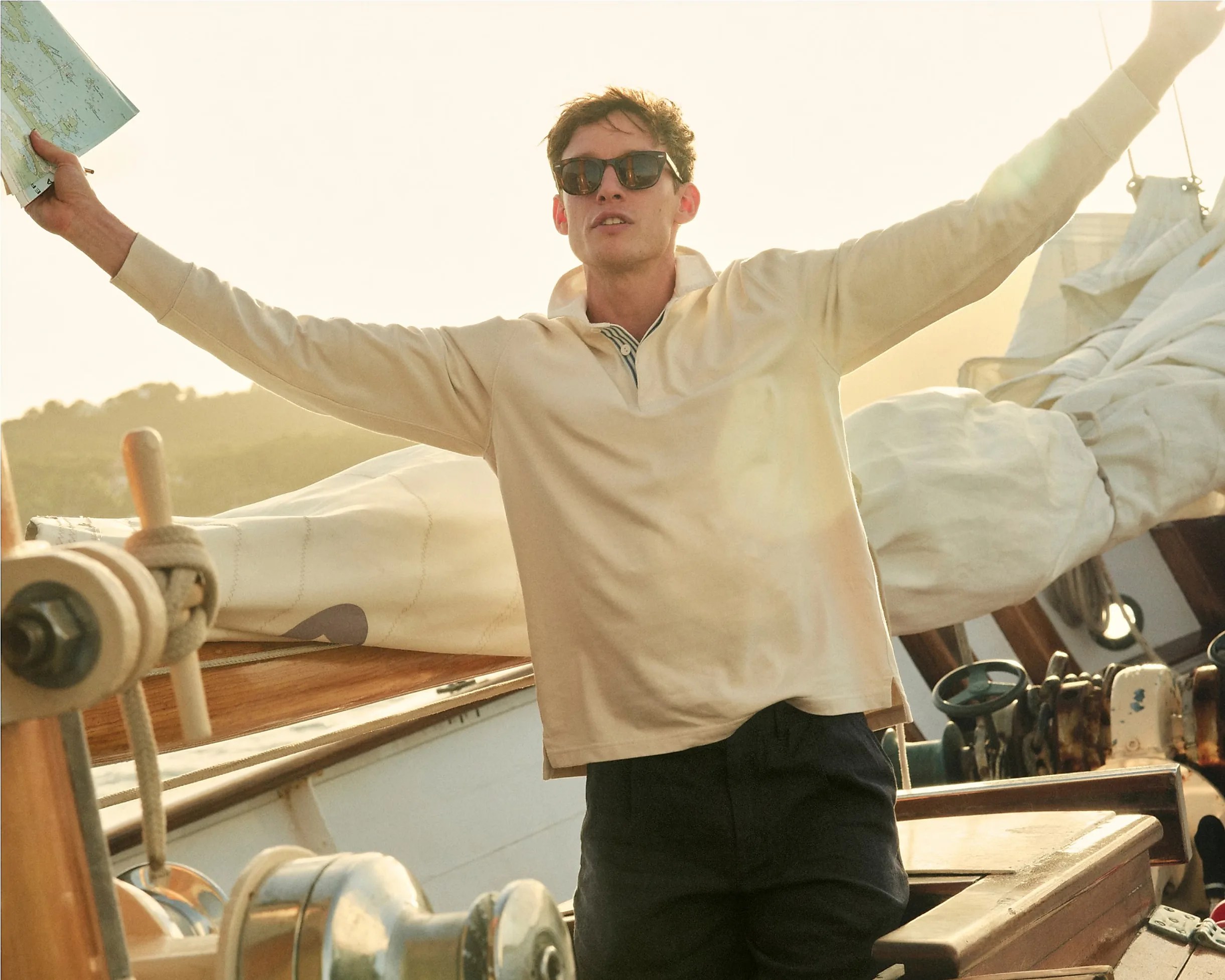 A young man wearing sunglasses and a light beige long-sleeve shirt stands on a sailboat with his arms outstretched. He holds a folded map in his left hand. The background shows sails and rigging of the boat, with a soft, warm light suggesting either sunrise or sunset.