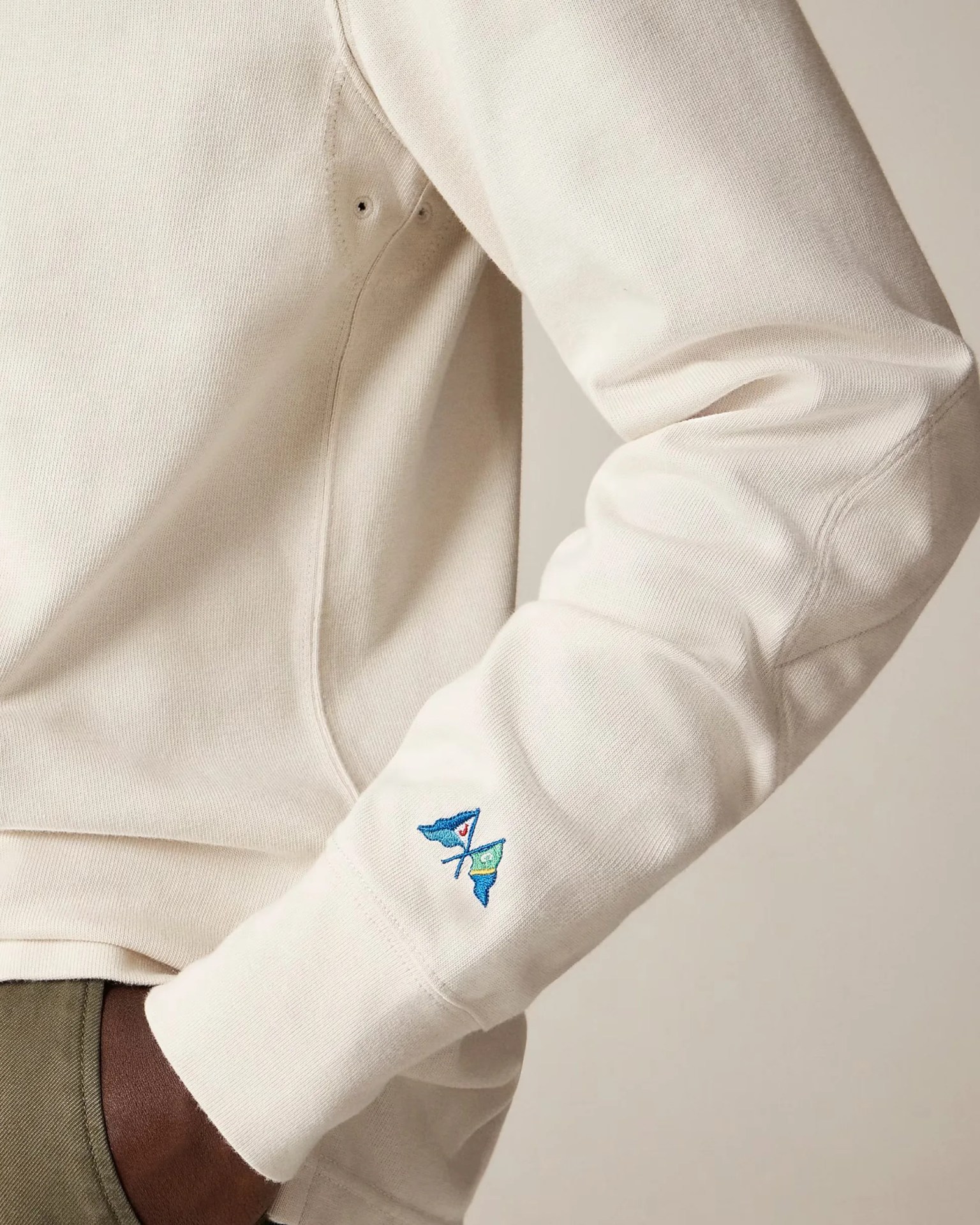 Cream-colored long sleeve shirt with a small embroidered design of two blue flags crossed on the lower sleeve near the wrist. The shirt has visible stitching and two small eyelets near the underarm area.