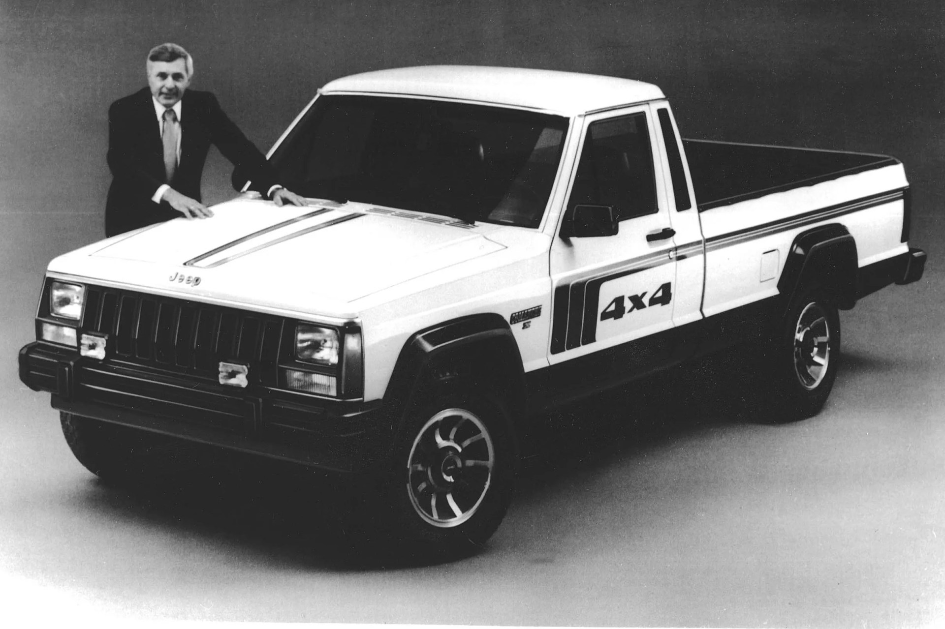 White Jeep 4x4 pickup truck with black trim and racing stripes on the hood and side, shown in a studio setting with a man in a suit standing behind it.