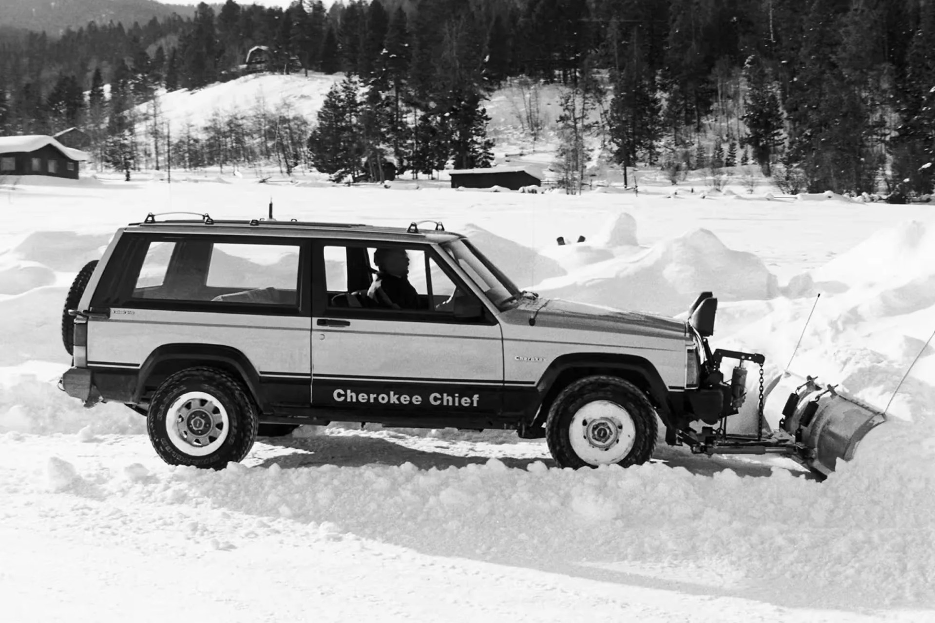 A Jeep Cherokee Chief equipped with a front-mounted snow plow is clearing snow on a snowy landscape. The vehicle is parked sideways, with a person visible inside the driver's seat. Snow-covered hills and trees are in the background.
