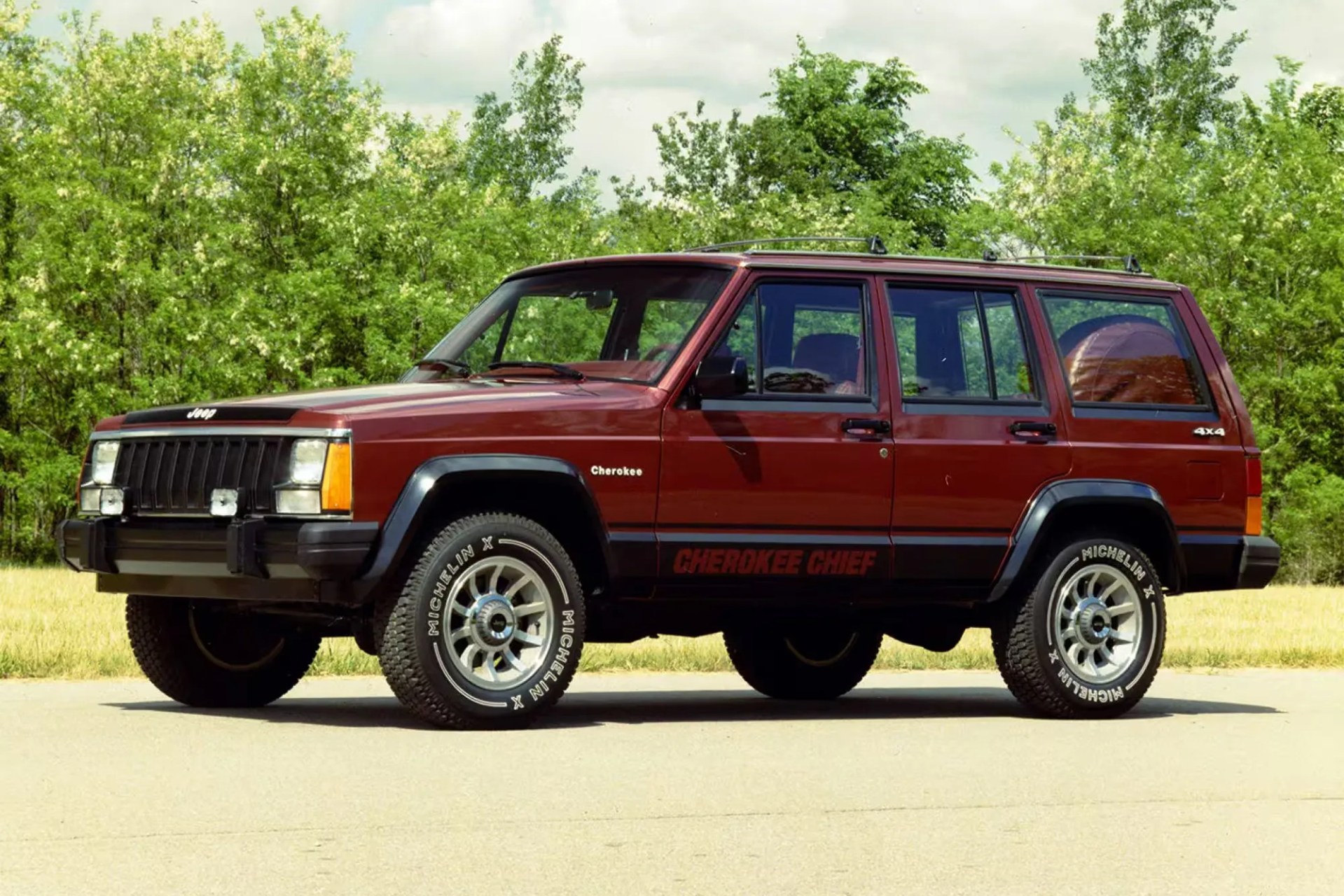 Maroon Jeep Cherokee Chief 4x4 SUV with silver alloy wheels and Michelin tires, parked on a paved surface with green trees in the background.