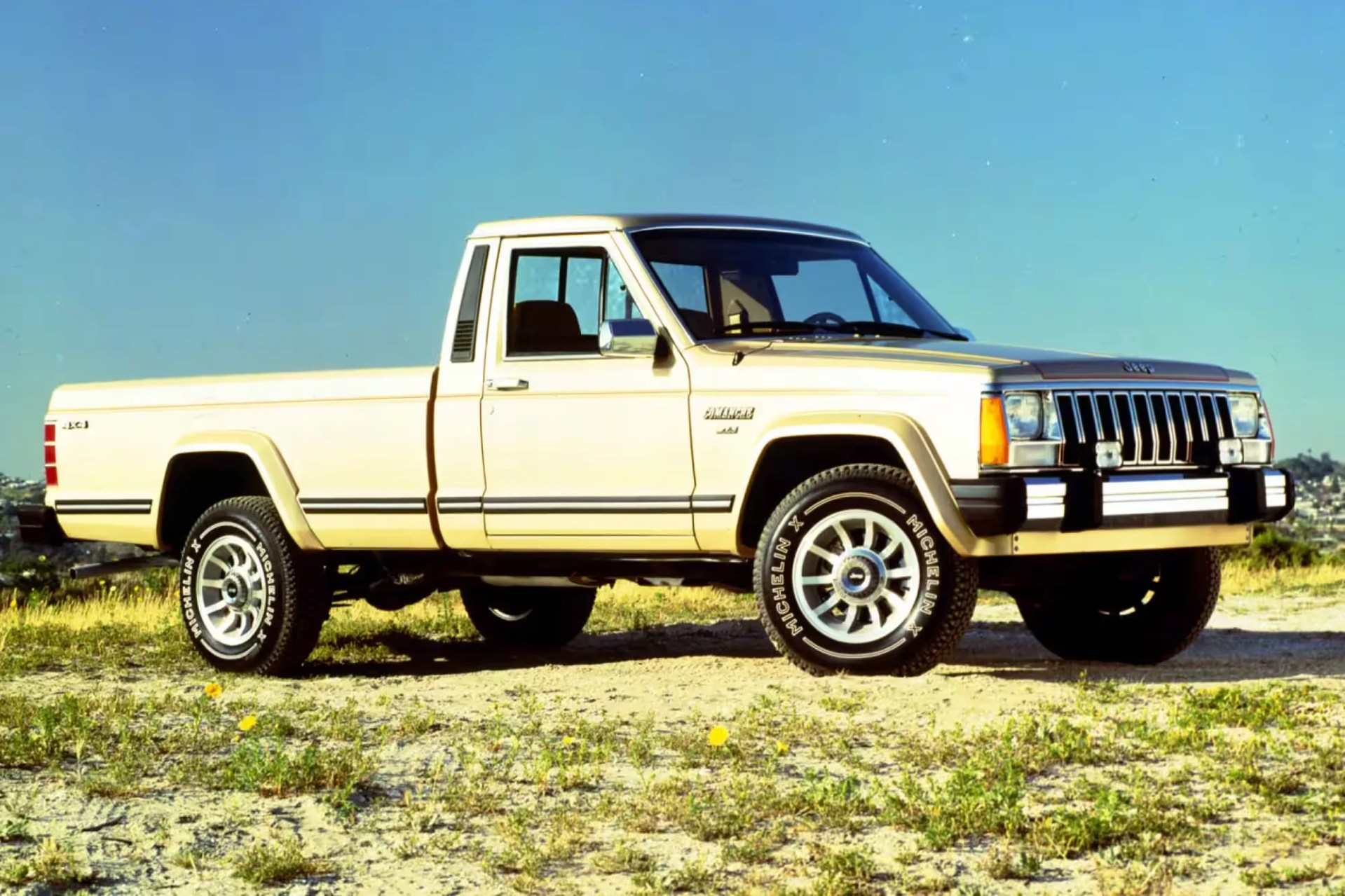 Beige vintage Jeep Comanche pickup truck parked on a dirt and grassy area under a clear blue sky. The truck has a single cab, chrome side mirrors, and Michelin tires with white lettering. The front grille features vertical slats and rectangular headlights.