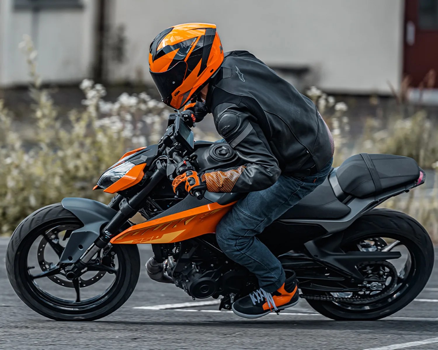 A person wearing an orange and black helmet, black leather jacket, orange gloves, and black and orange shoes is riding an orange and black sport motorcycle on a paved surface. The rider is leaning forward in a racing posture. The background is blurred with some greenery and a building.