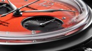 Close-up of a kurono tokyo watch face with an orange dial, featuring silver hour and minute hands, a black subdial with white markings, and silver hour markers. The watch has a polished metal bezel and a curved crystal. The number "12" is prominently displayed at the top, and some text is partially visible on the dial.