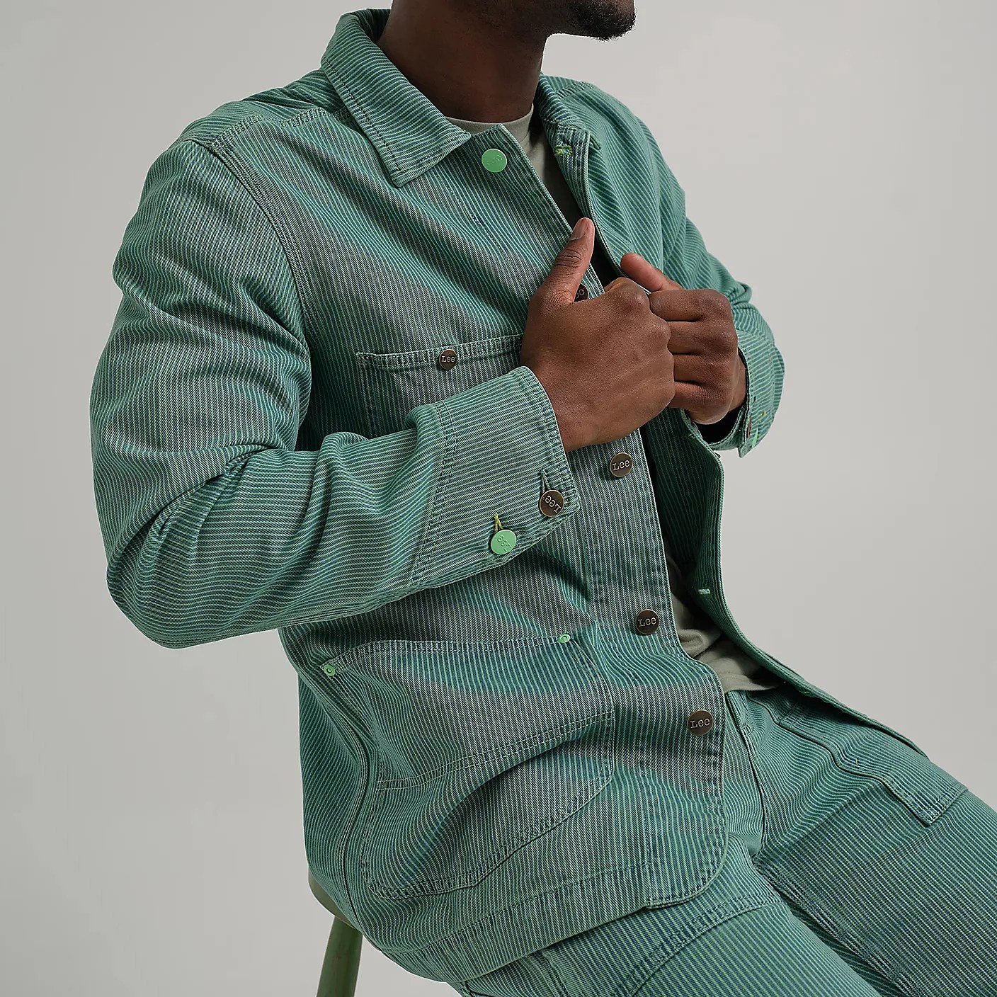 Green and white pinstriped denim jacket with matching pants, featuring metal buttons labeled "Lee" and large front pockets. The jacket has a collar and buttoned cuffs. The person wearing the outfit is seated and holding the jacket closed.