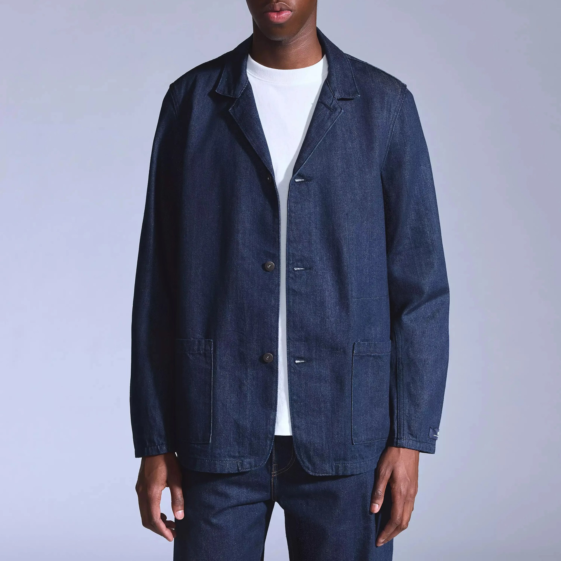 Dark blue denim jacket with a notched collar, button front closure, and two front patch pockets, worn over a white t-shirt.