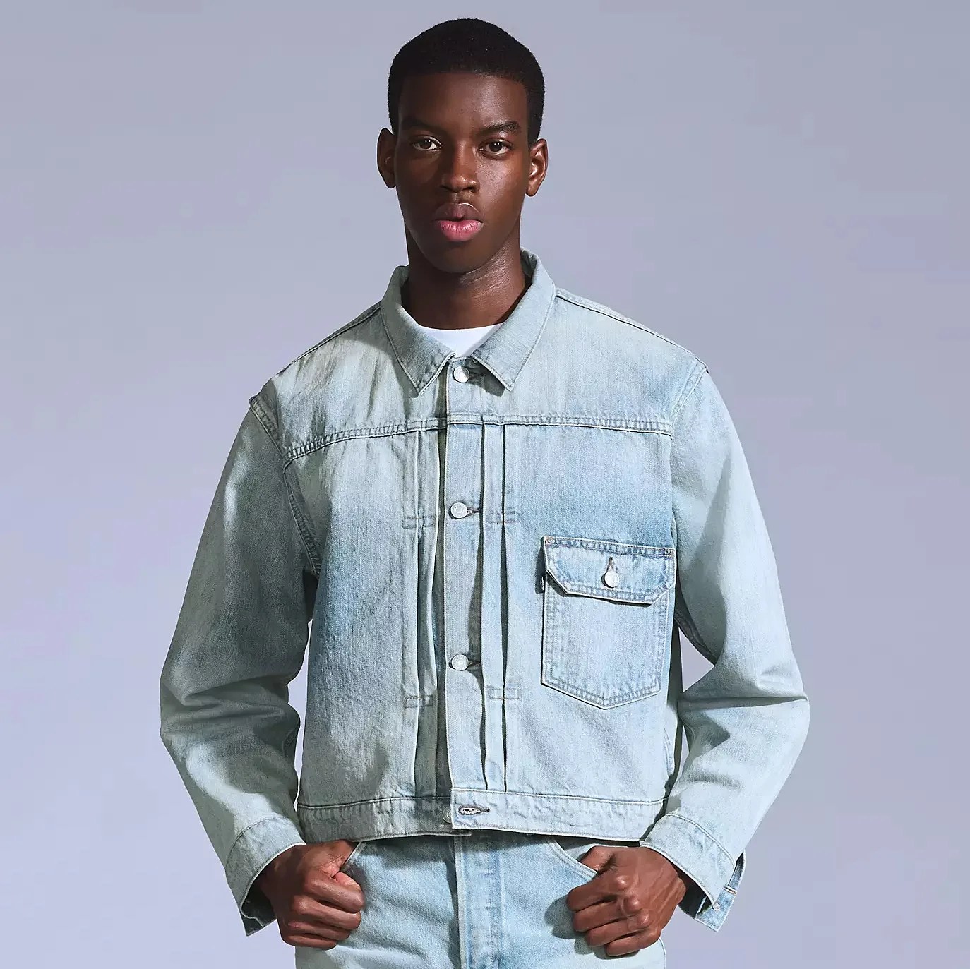 Light blue denim jacket with a button-up front and a single flap pocket on the left chest, worn by a person with hands tucked into the front pockets of matching light blue denim pants.