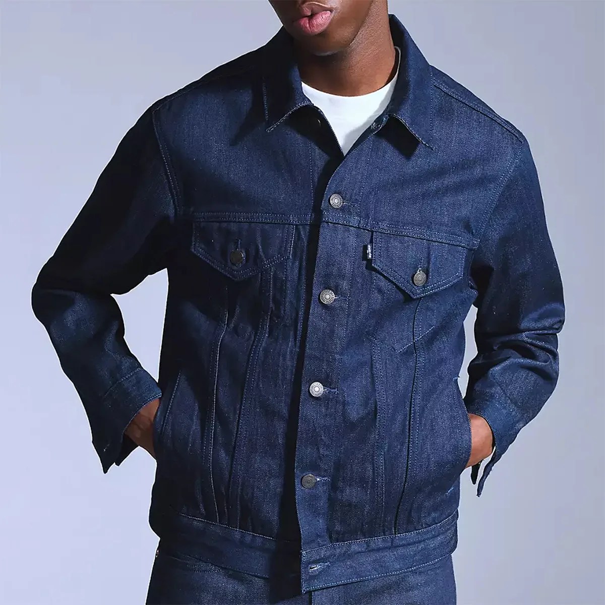 Dark blue denim jacket with button closure, two chest flap pockets with buttons, and side pockets. The jacket has a classic collar and visible stitching details. The person wearing it has their hands in the side pockets.