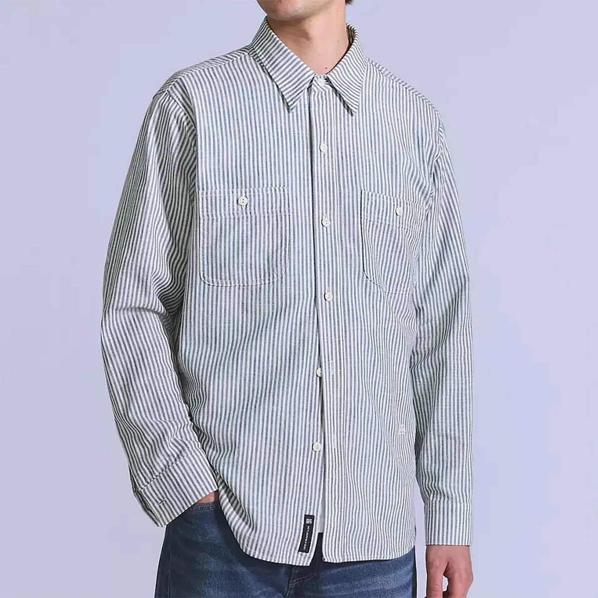 Long-sleeve button-up shirt with vertical white and gray stripes, featuring a pointed collar and two buttoned chest pockets. The shirt is worn untucked over blue jeans.